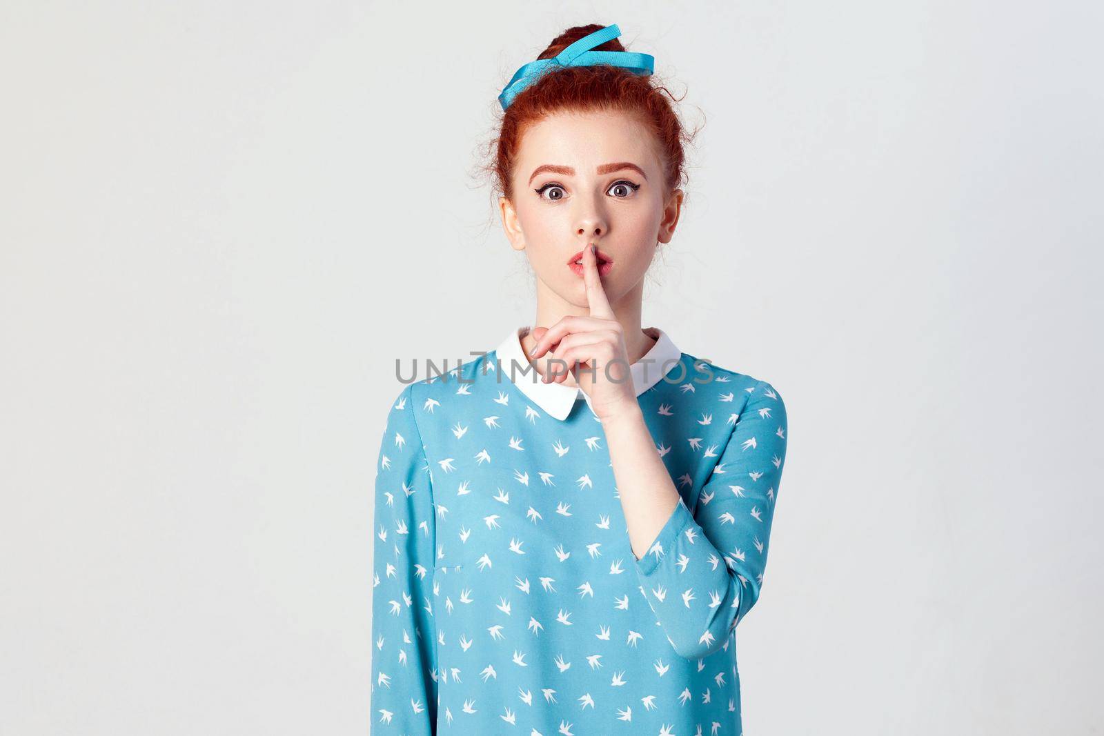 The beautiful young redhead girl, holding index finger at lips, raising brows Isolated studio shot on gray background