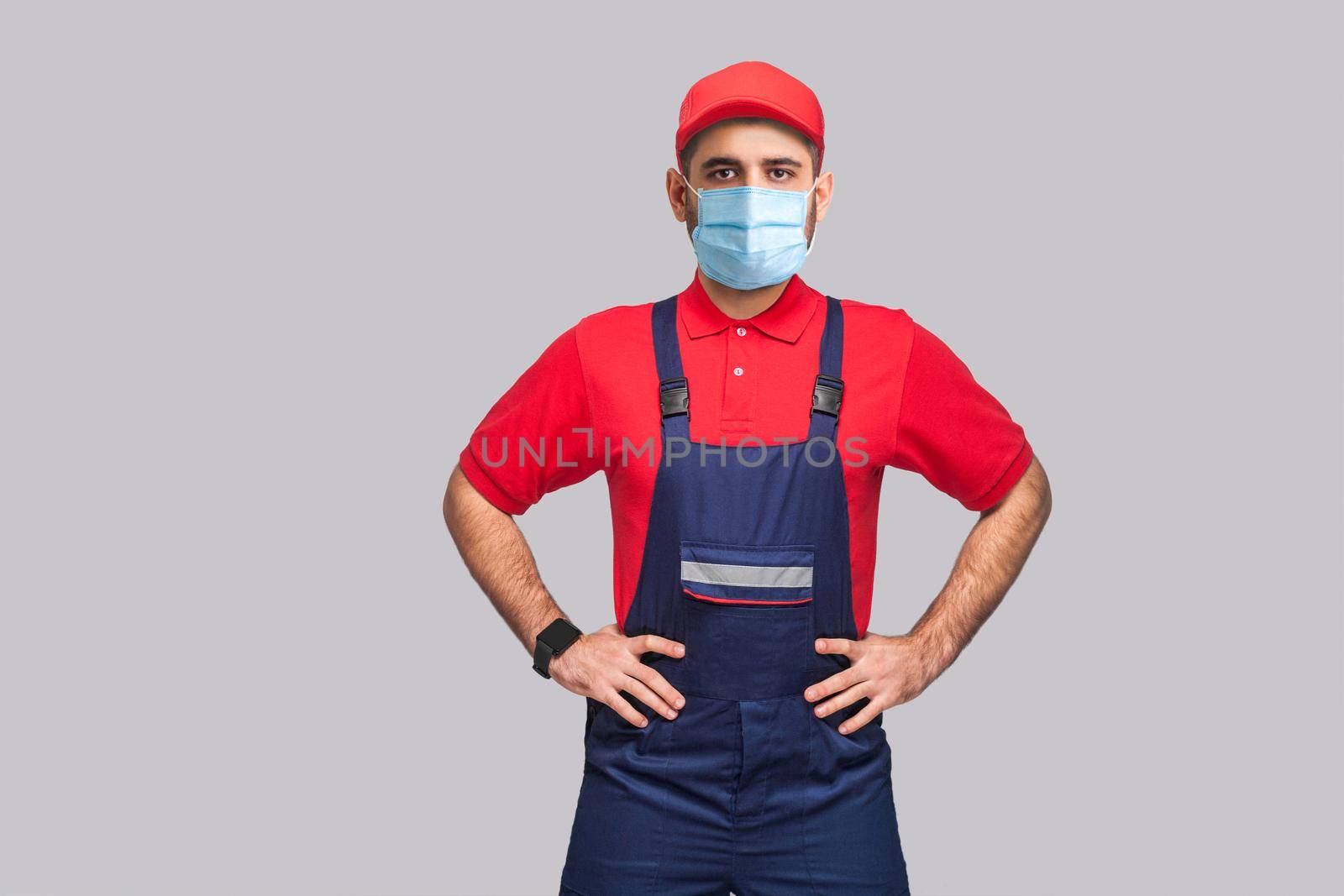 Portrait of young man with surgical medical mask in blue overall, red t-shirt and cap standing and holding hands on waist and looking at camera, indoor, studio shot, isolated on gray background