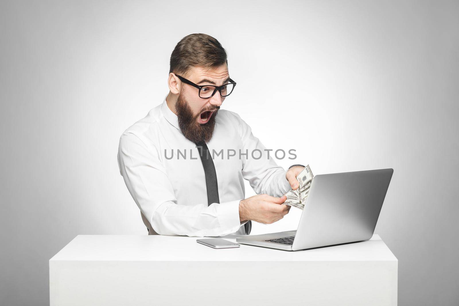 Portrait of emotional shocked businessman in white shirt are sitting in office holding cash with suprised faced and opened mouth looking to the laptop. indoor studio shot isolated on grey background.