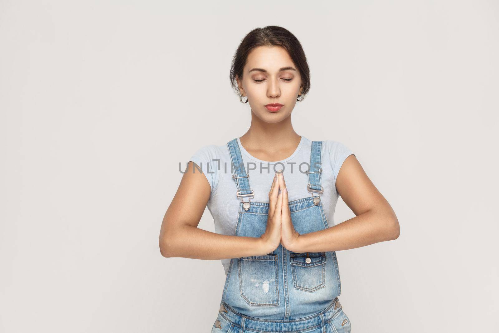 Beautiful young caucasian female, holding hands in namaste or prayer, keeping eyes closed while practising yoga and meditating at home alone. Studio shot on gray background.