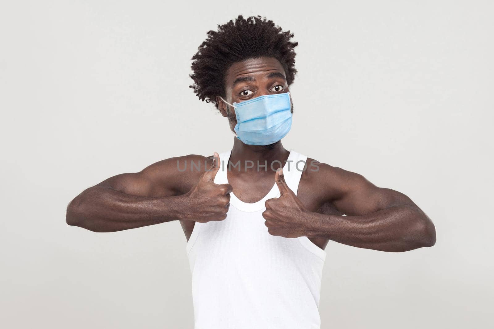 wow, I like it. Portrait of satisfied young man wearing white shirt with surgical medical mask standing, looking at camera and showing thumbs up. indoor studio shot isolated on gray background.