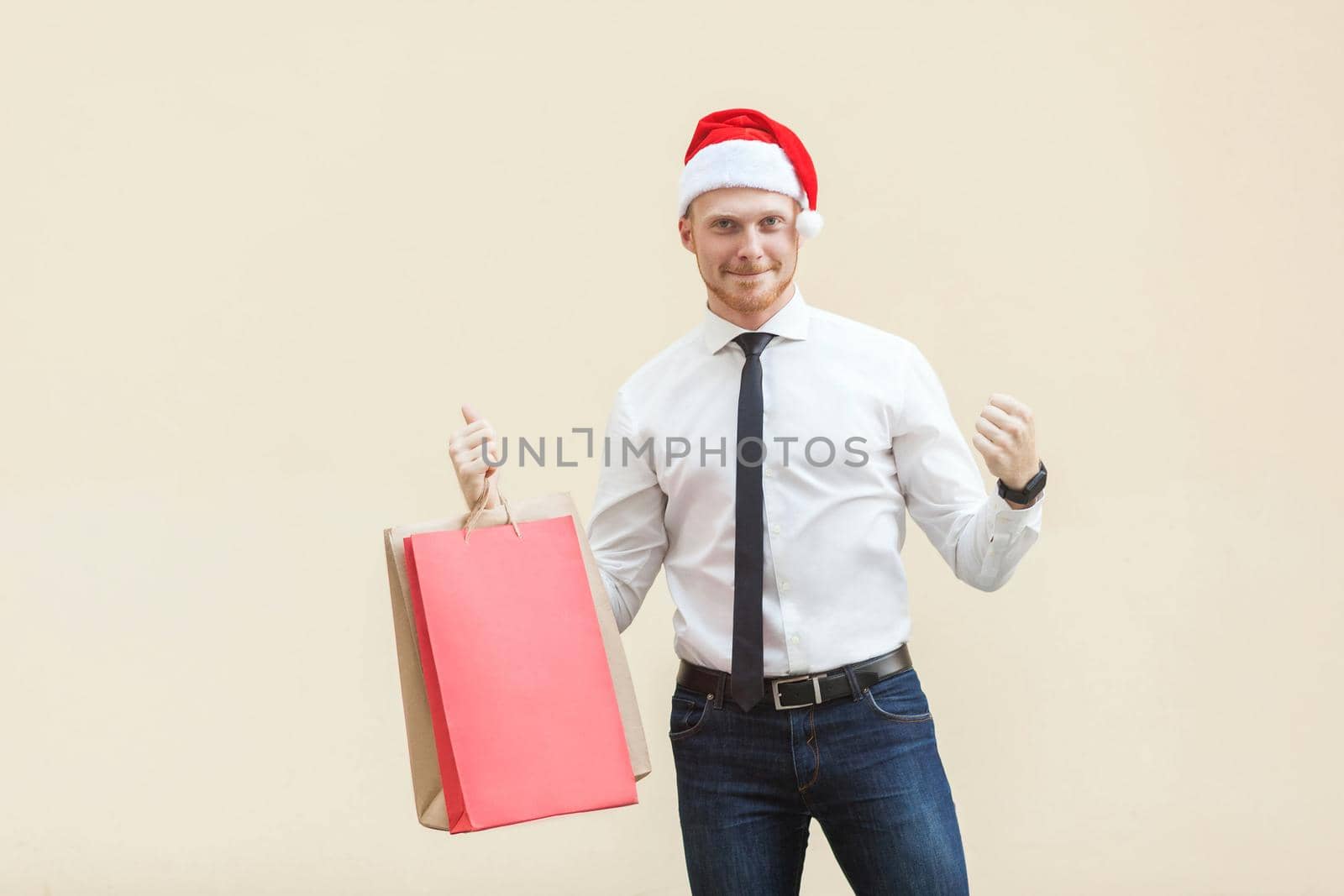 Buy for best price in winter. Young adult and beard man in red santa hat ,happiness looking at camera. Indoor, studio shot on light orange background