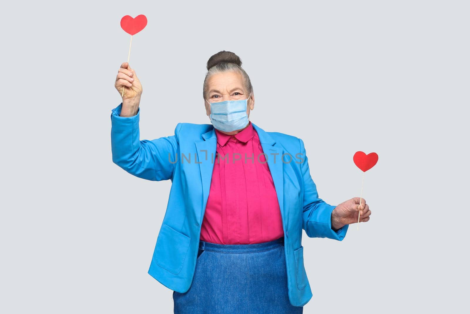 happy aged with surgical medical mask woman holding two red heart sticker. Portrait of beautiful happy and satisfied grandmother in light blue suit. indoor studio shot, isolated on gray background