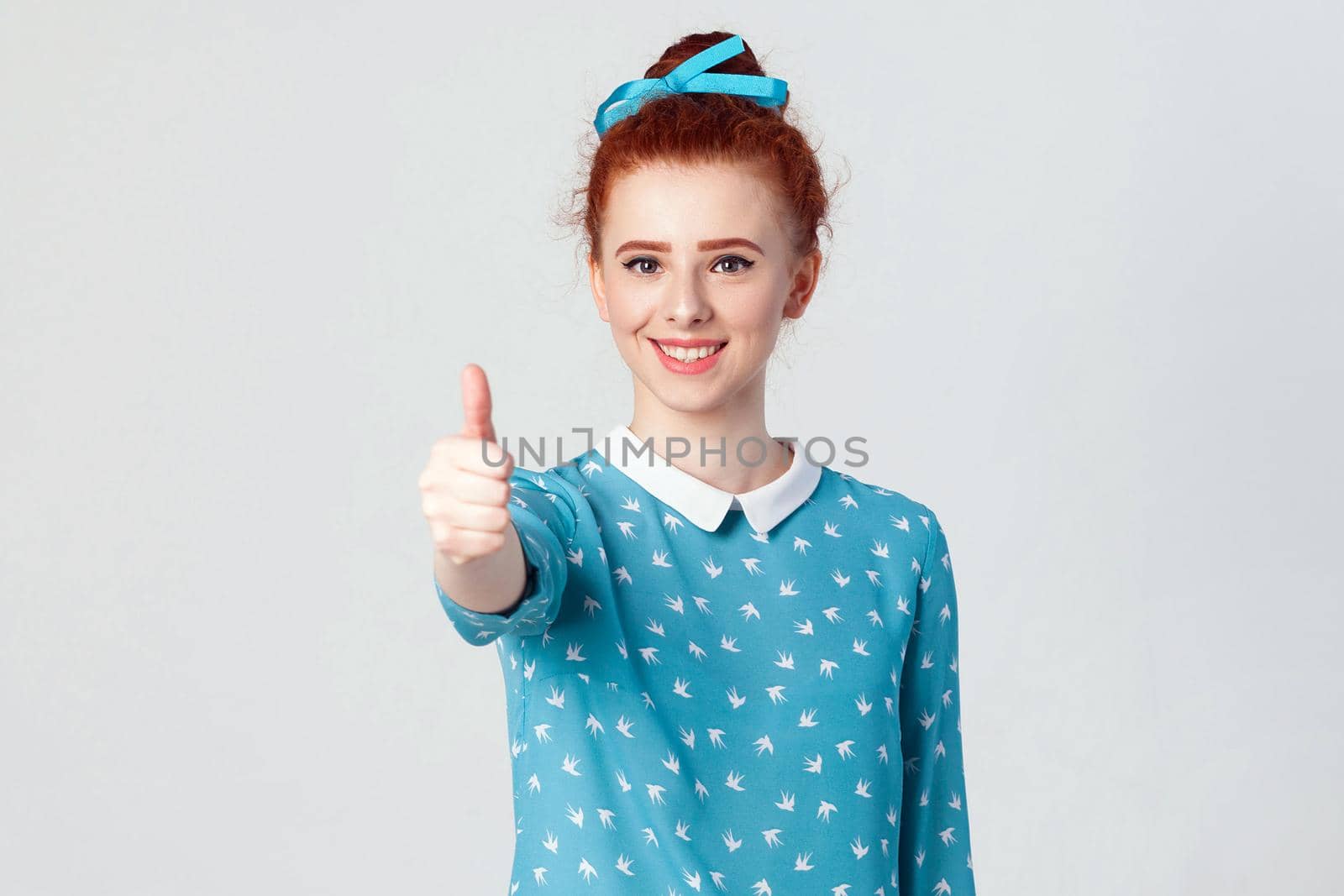 Young happy cheerful redhead girl showing thumb up and toothy smile. Isolated studio shot on gray background