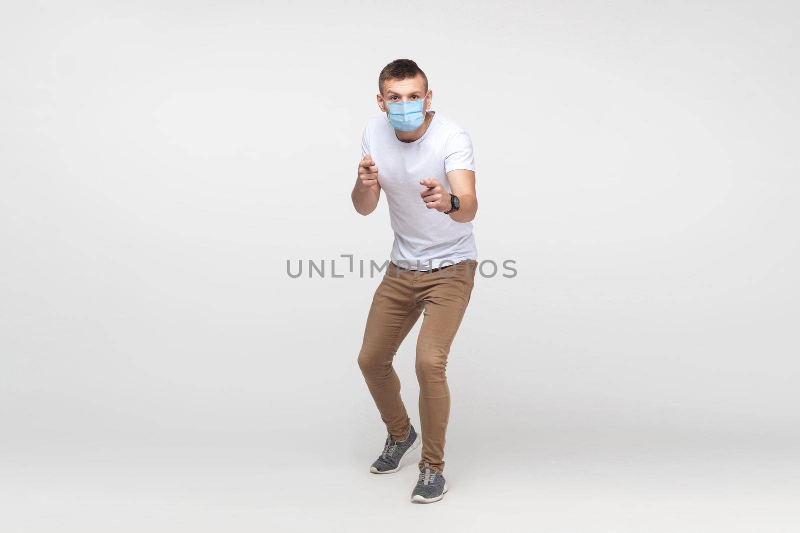 Hey you. Full length portrait of serious young man in white shirt with surgical medical mask standing, looking and pointing at camera with serious face. indoor studio shot, isolated on gray background