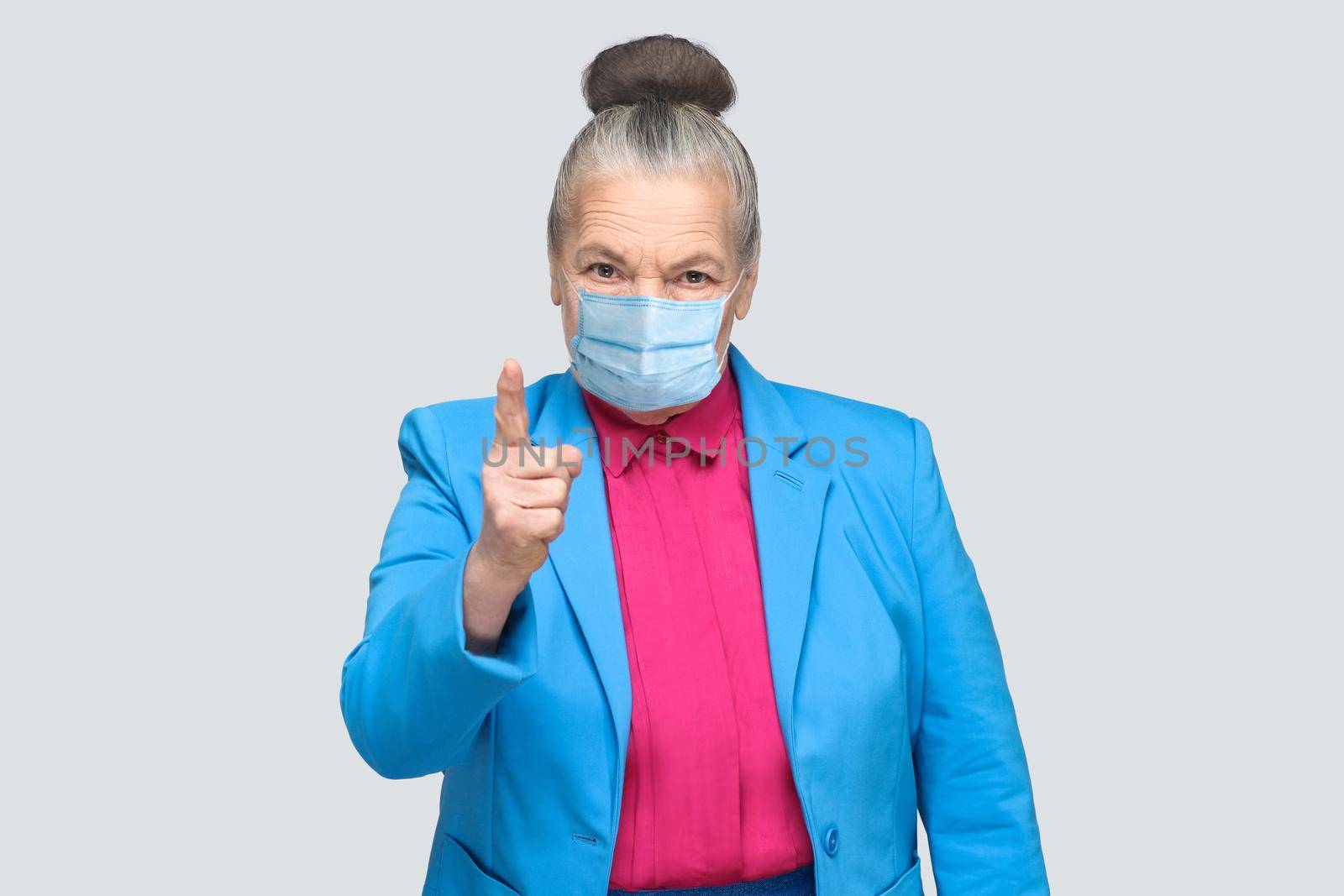 Angry aged woman with surgical medical mask warning you. Grandmother with light blue suit and pink shirt standing with collected bun gray hair. indoor studio shot, isolated on gray background