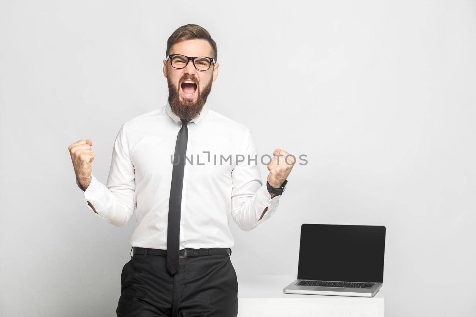 Yeah! Portrait of handsome happy bearded young businessman in white shirt and black tie are standing in office are triumphing with open mouth. Isolated, studio shot, indoor, gray background