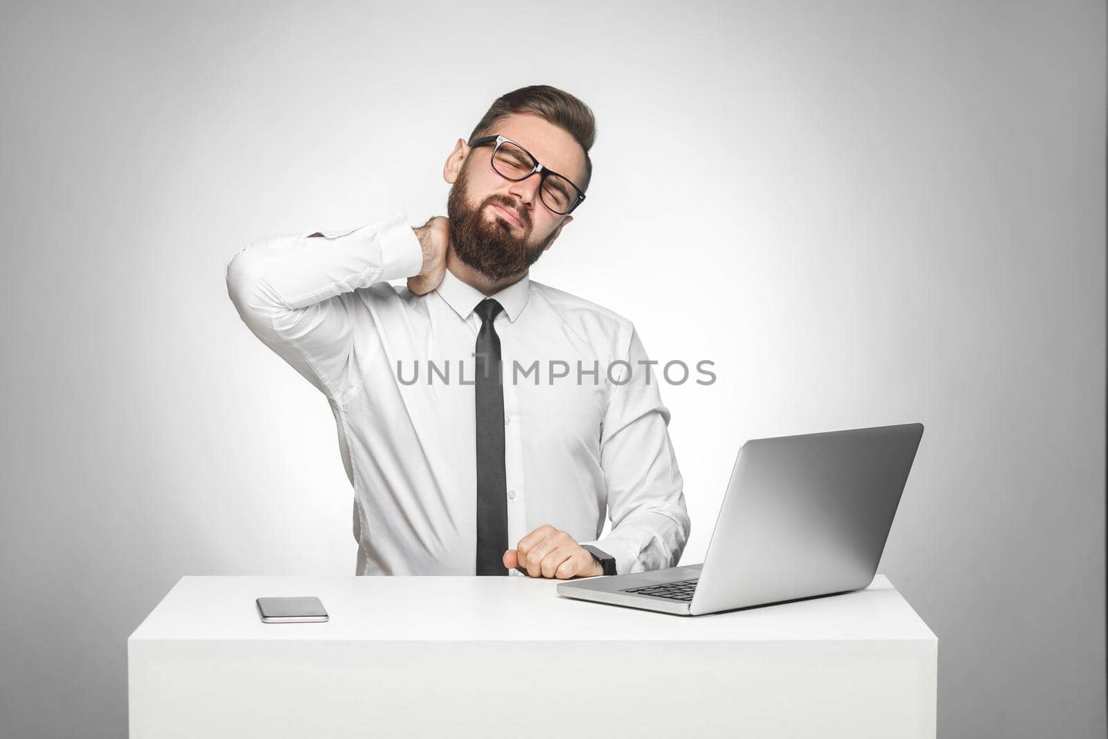 Portrait of unhealphy upset tired young boss in white shirt and black tie are sitting in office and have strong pain in neck, holding hand on neck. Studio shot, isolated, gray background, indoor