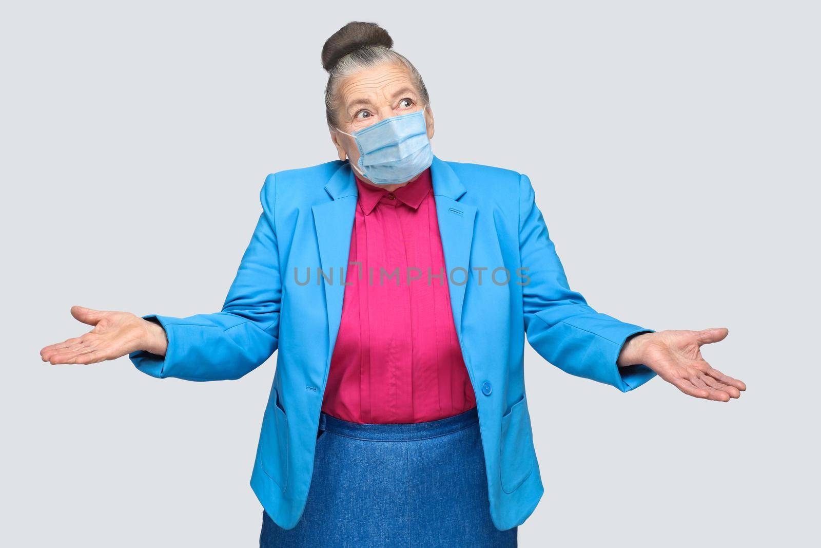 Puzzled or confused old woman with surgical medical mask, raised arms. grandmother with light blue suit and pink shirt standing with collected bun gray hair. indoor shot, isolated on gray background