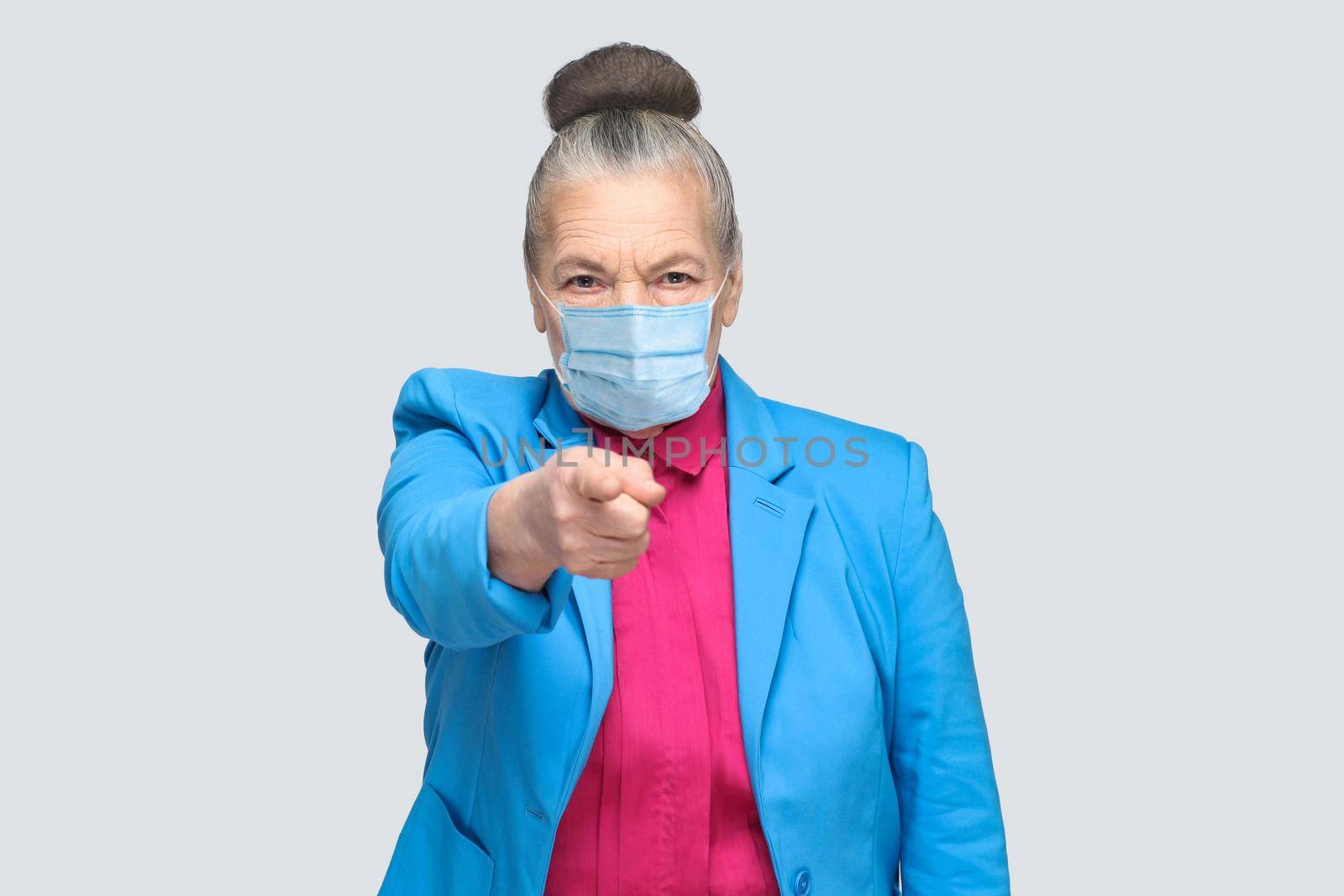 Hey you! Woman with surgical medical mask pointing at camera finger with serious face. Portrait of expressive grandmother standing with collected hair. indoor studio shot, isolated on gray background