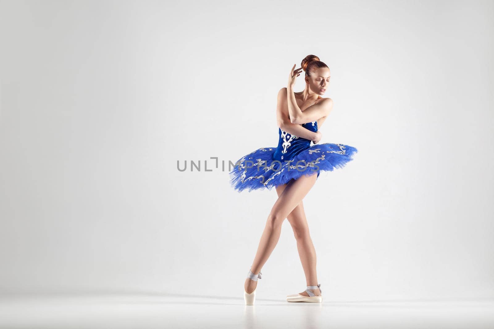 Young beautiful ballerina with bun collected hair wearing blue dress and pointe shoes dancing gracefully isolated on white background. indoor, studio shot.