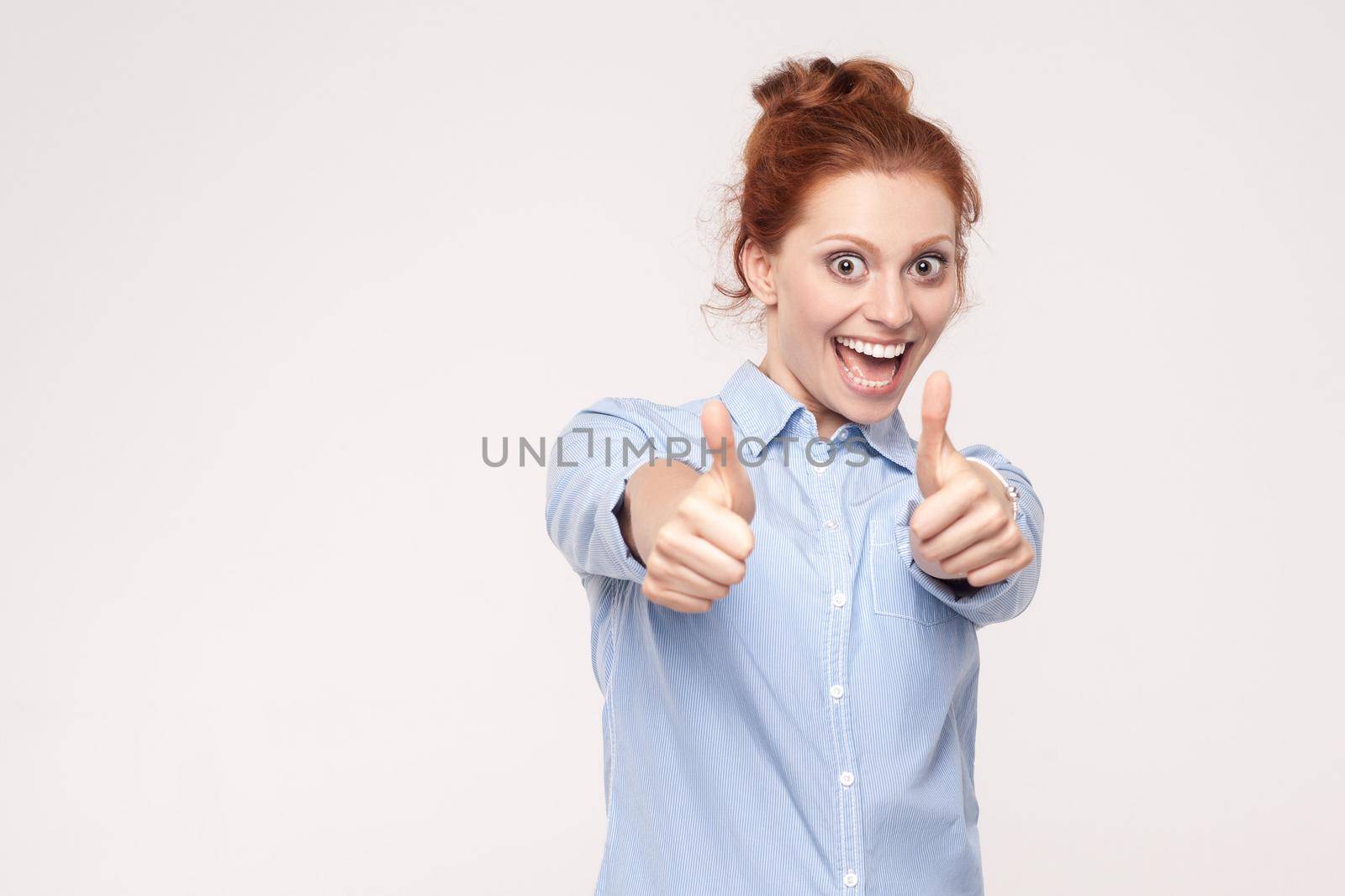 Happy cheerful redhead woman showing thumb up and toothy smile. Isolated studio shot on gray background