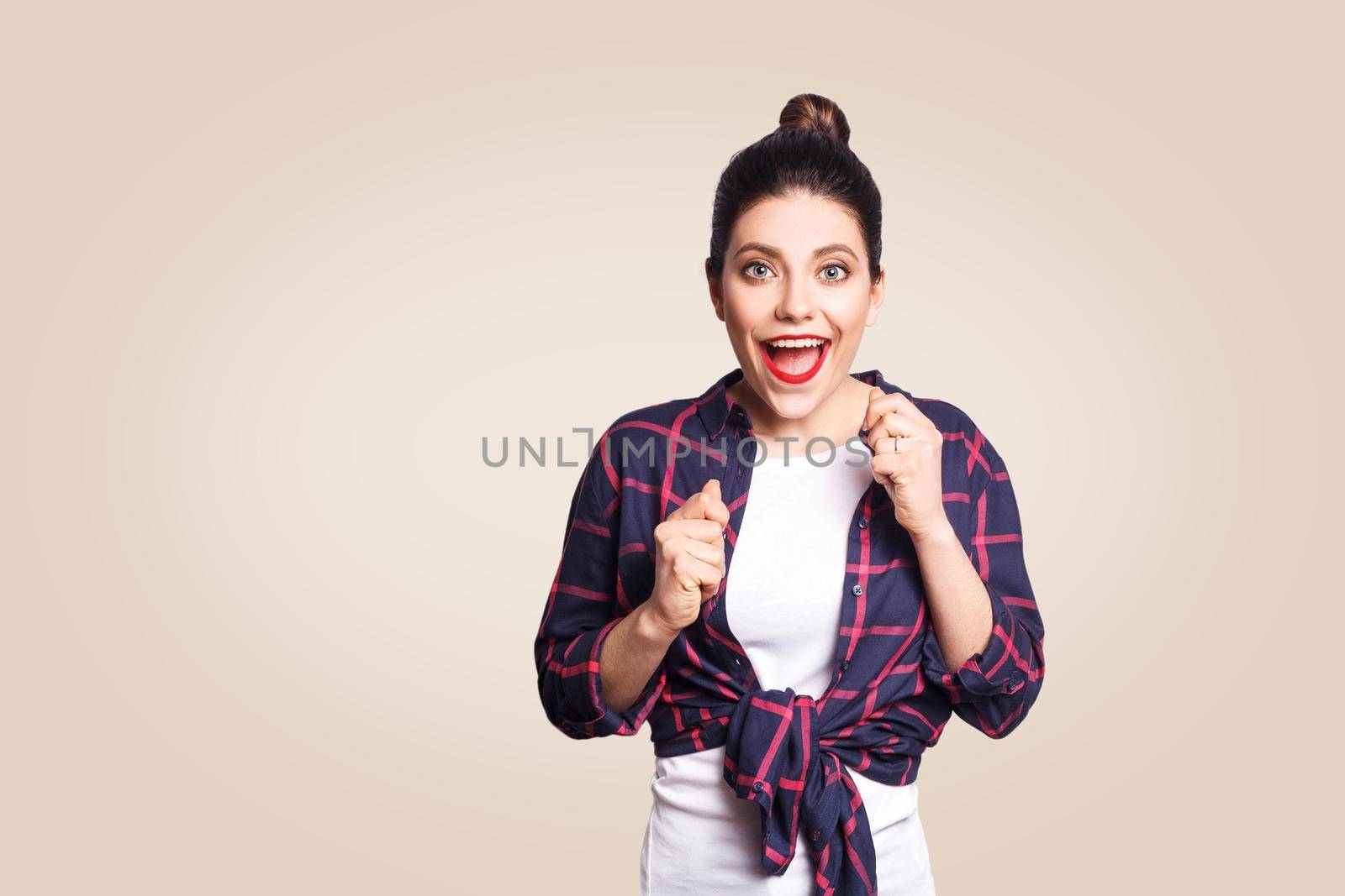 Portrait of pretty girl having winning and happy facial expression, exclaiming with joy, keeping hands in fists and mouth wide open, cheering, celebrating success or positive news, posing indoors.