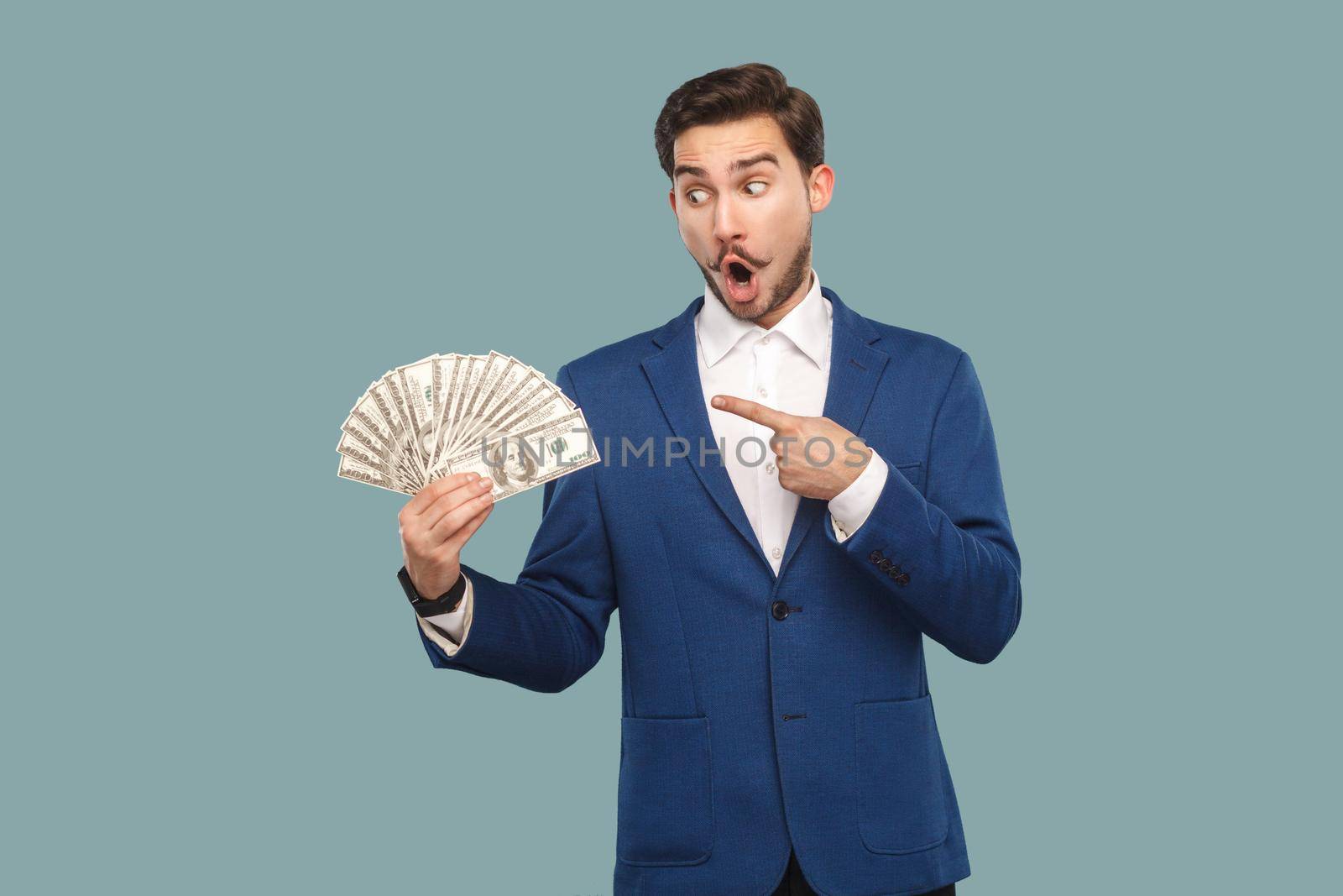 Handsome wondered businessman in blue jacket standing and holding many dollars in hand, pointing and looking at money with amazed surprised face. Indoor, studio shot isolated on light blue background.