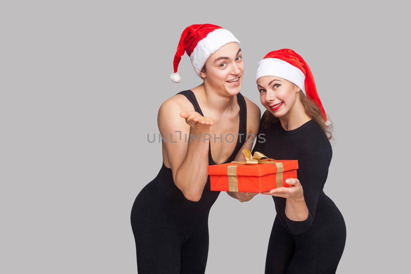 It's gift box for you! couple in christmas hat standing and sharing gift box and looking at camera. Indoor, studio shot, isolated on gray background