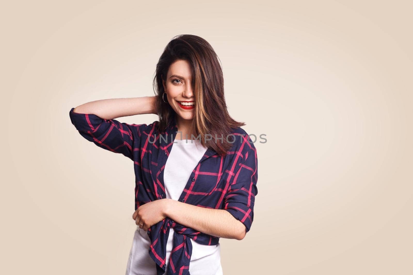 Portrait of beautiful happy girl in casual style with makeup and black bun hairstyle looking at camera with toothy smile and touching her hair. studio shot on beige background.