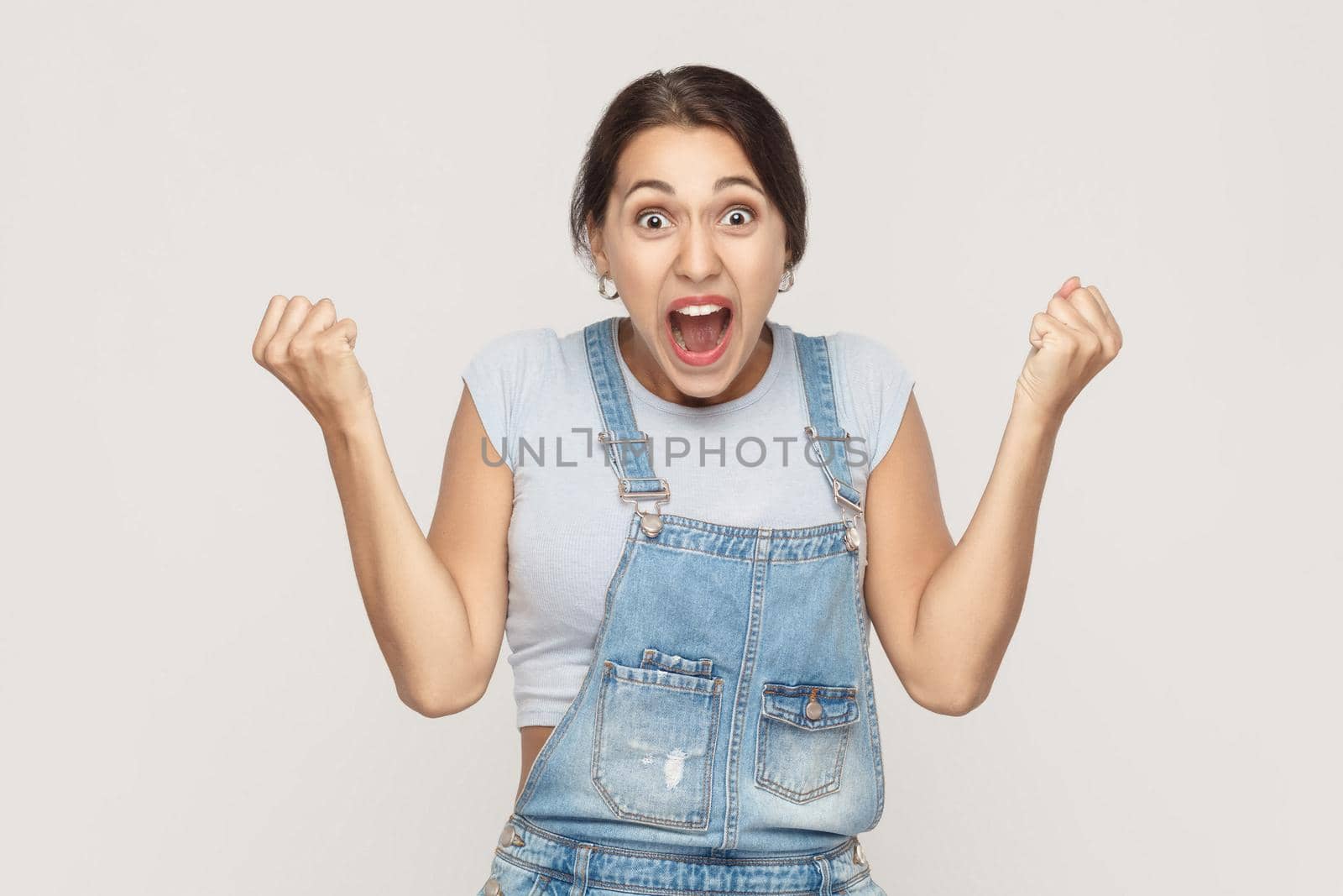 Happiness woman in denim overalls rejoicing for his success. Isolated on gray background. Indoor, studio shot