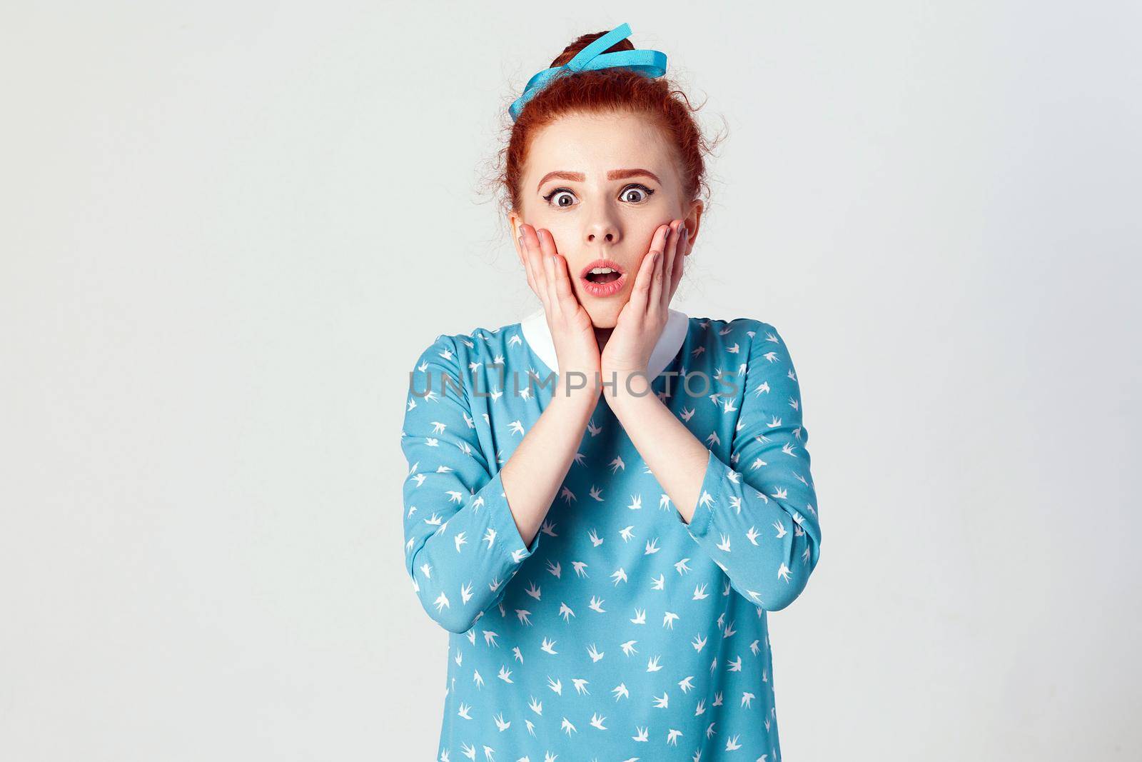 Human face expressions and emotions. Redhead young girl screaming with shock, holding hands on her cheeks. Isolated studio shot on gray background.