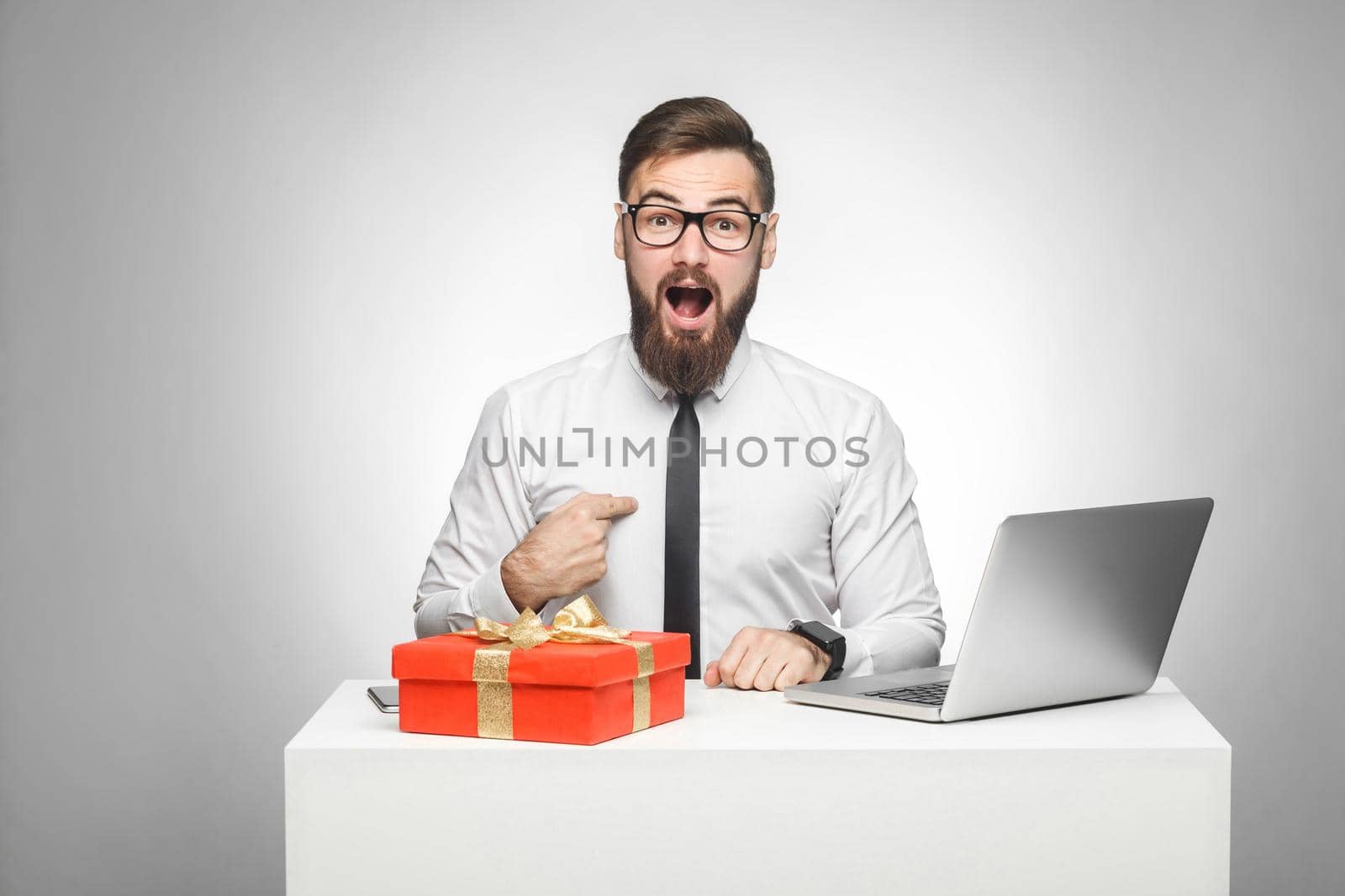 is it for me? shocked young man in white shirt and black tie are sitting in office and pointing finger to himself with shocked face, looking at camera. Indoor, isolated,studio shot,gray background