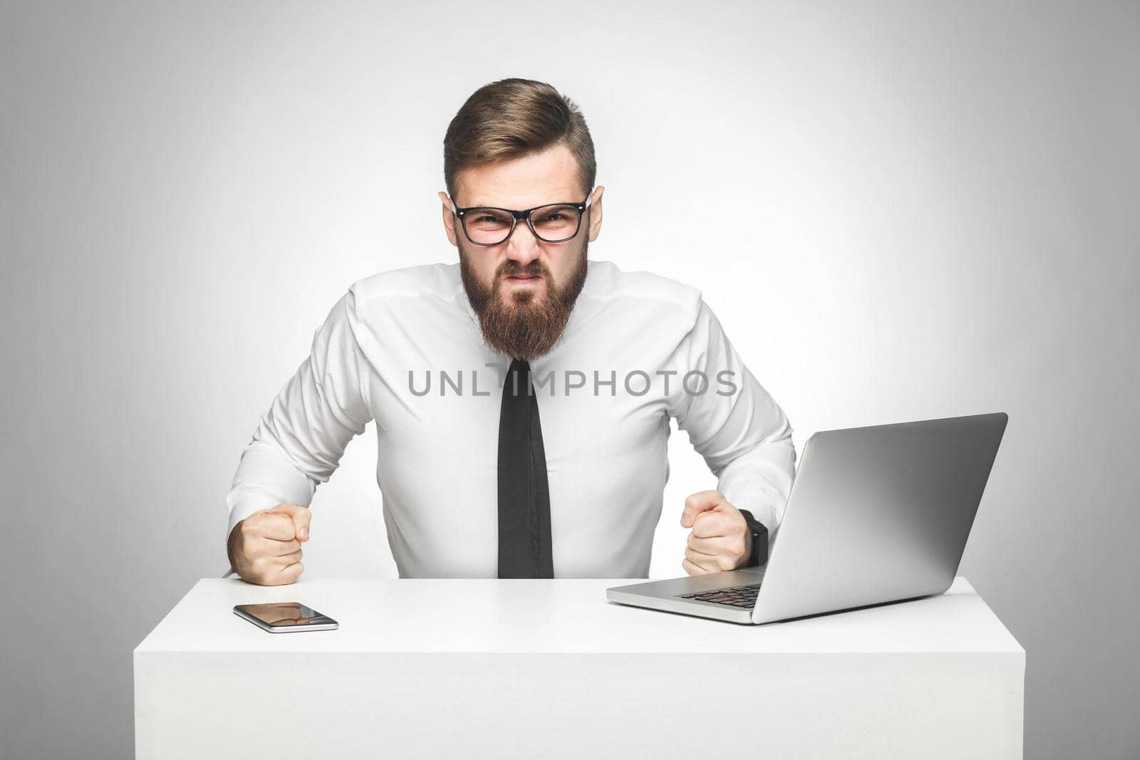 Portrait of aggressive angry young boss in white shirt and black tie are sitting in office and having bad mood,punch the table and clenching teeth. Indoor, studio shot, isolated, gray backgroud