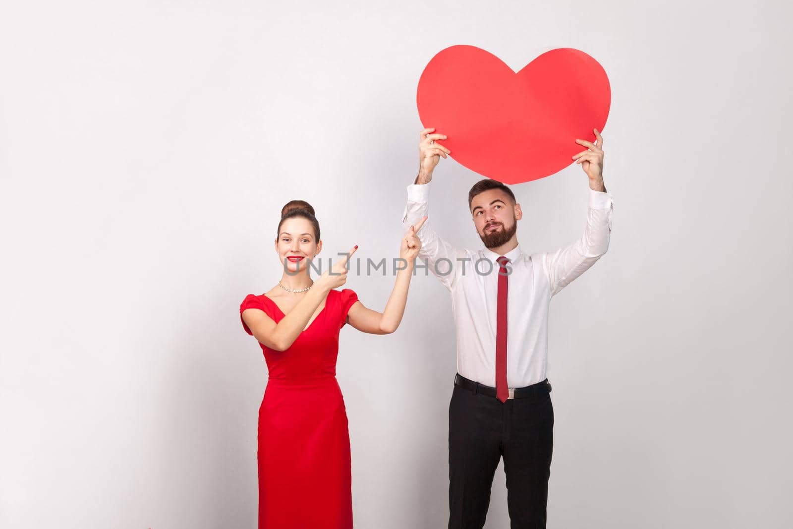 Woman pointing finger at big heart, man looking at heart. Indoor, studio shot, isolated on gray background