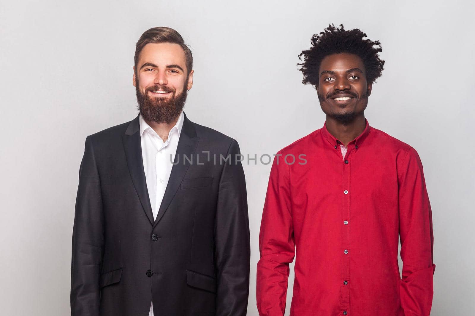 Two partners to business, standing near each other, looking at camera and toothy smiling. Studio shot, gray background