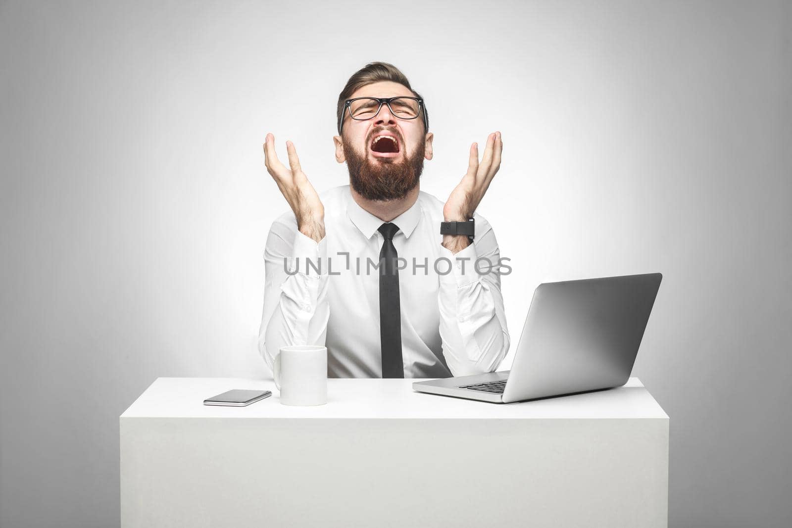 No way! Portrait of emotional scared young manager in white shirt and black tie are sitting in office and screaming and crying cause made big mistake with raised arms and stressed face. Studio shot