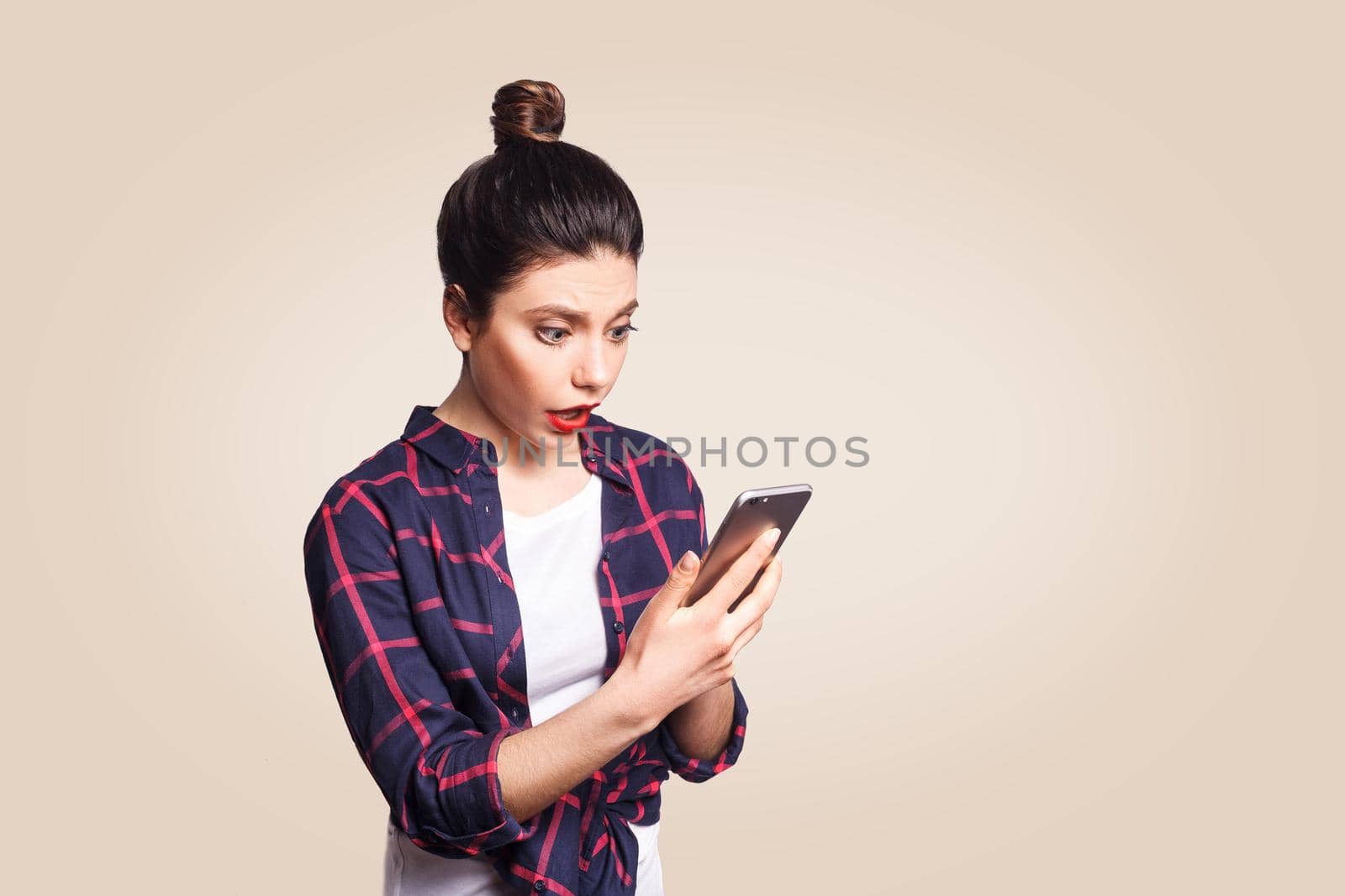 Young beautiful woman in casual style holding phone and surprised. looking at camera with shock. studio shot on beige background.