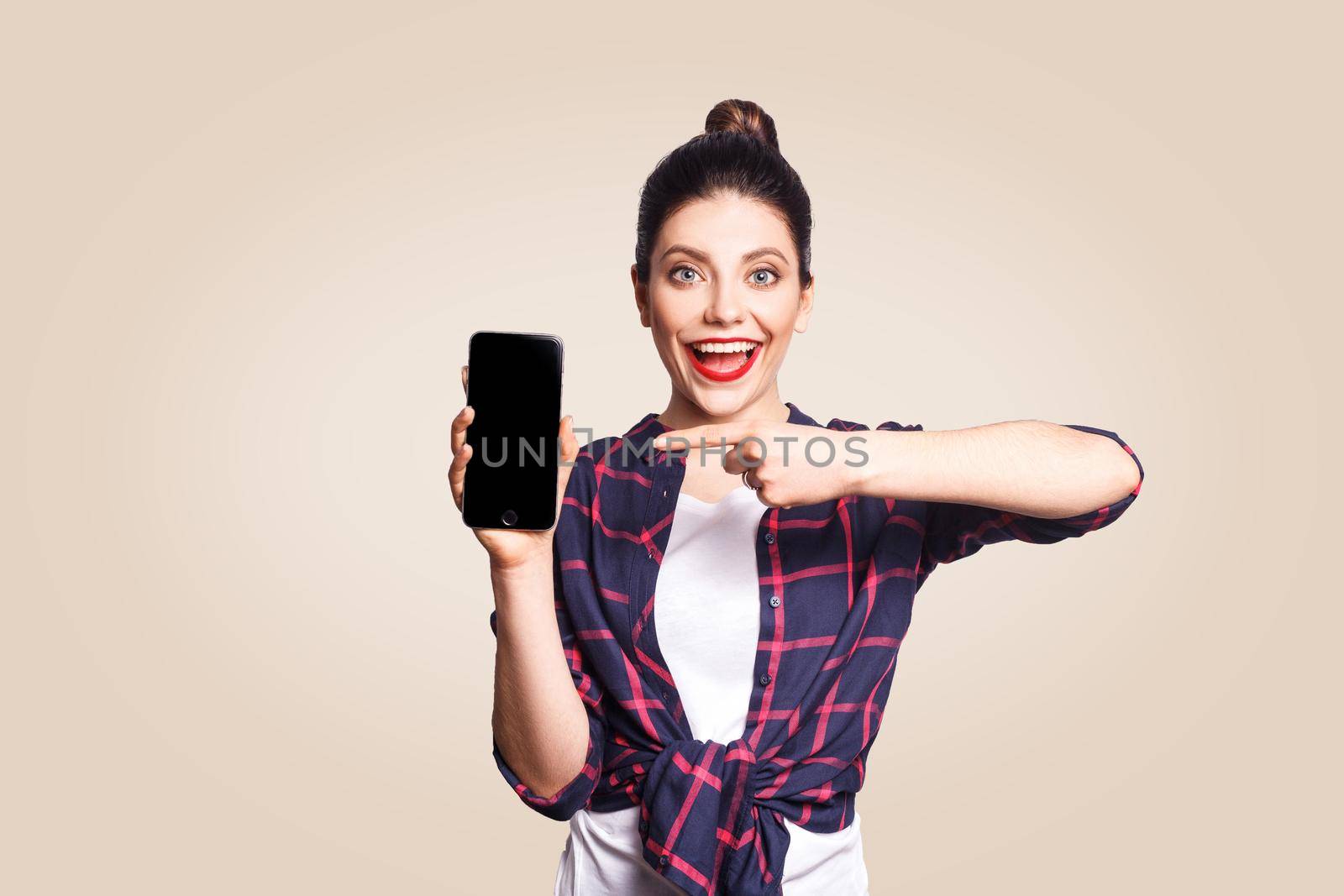 Young beautiful woman in casual style holding phone looking at camera and showing phone display with finger. studio shot on beige background.