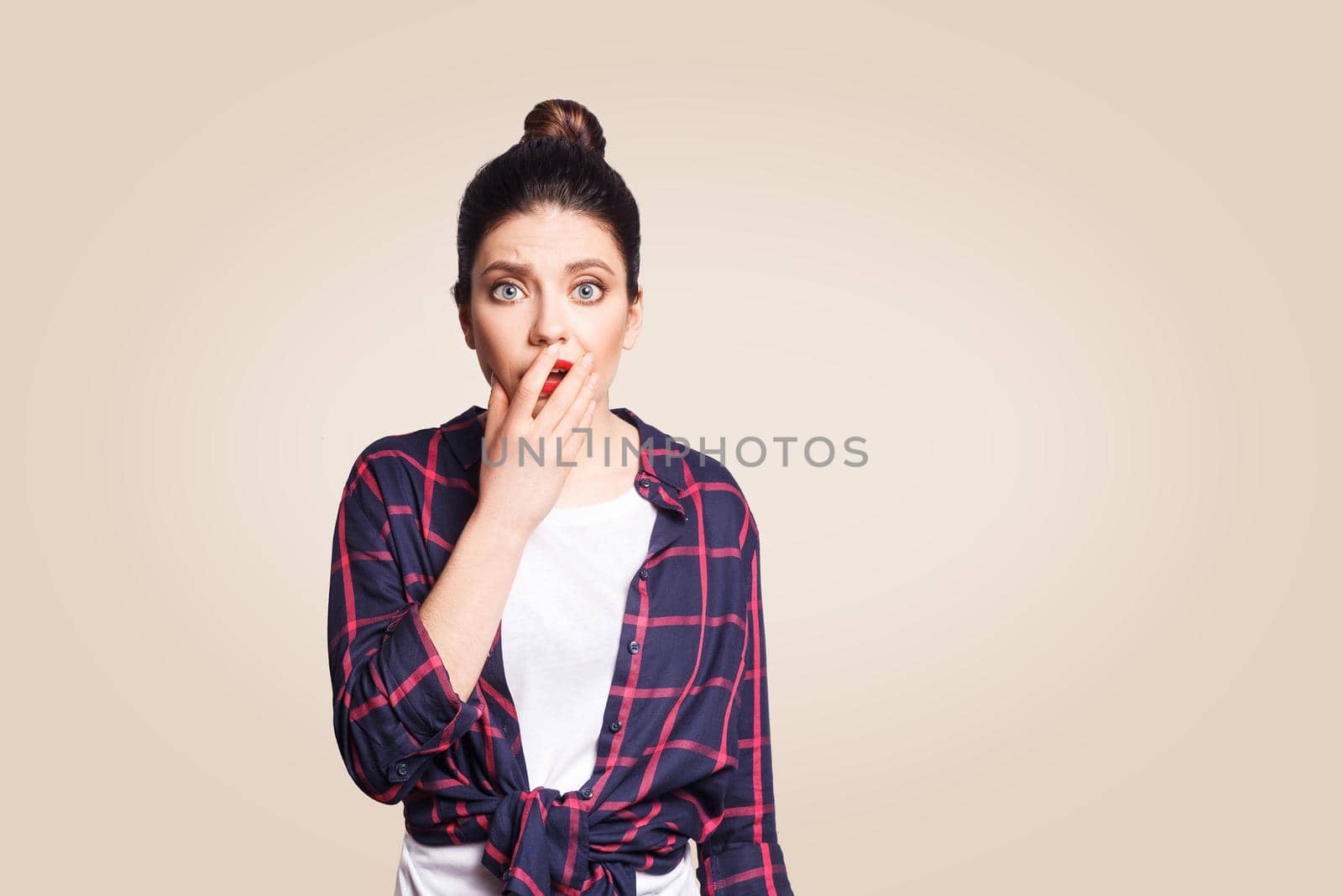 Bad news. Frightened startled young woman raising eyebrows, keeping hand on her cheek and staring at camera, having shocked scared look. studio shot on beige background.