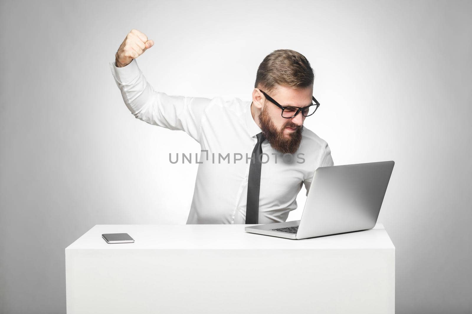 Portrait of aggressive unhappy businessman sitting in office and having bad mood are ready to punch a worker through a webcam with fist and angry face. indoor studio shot, isolated on grey background