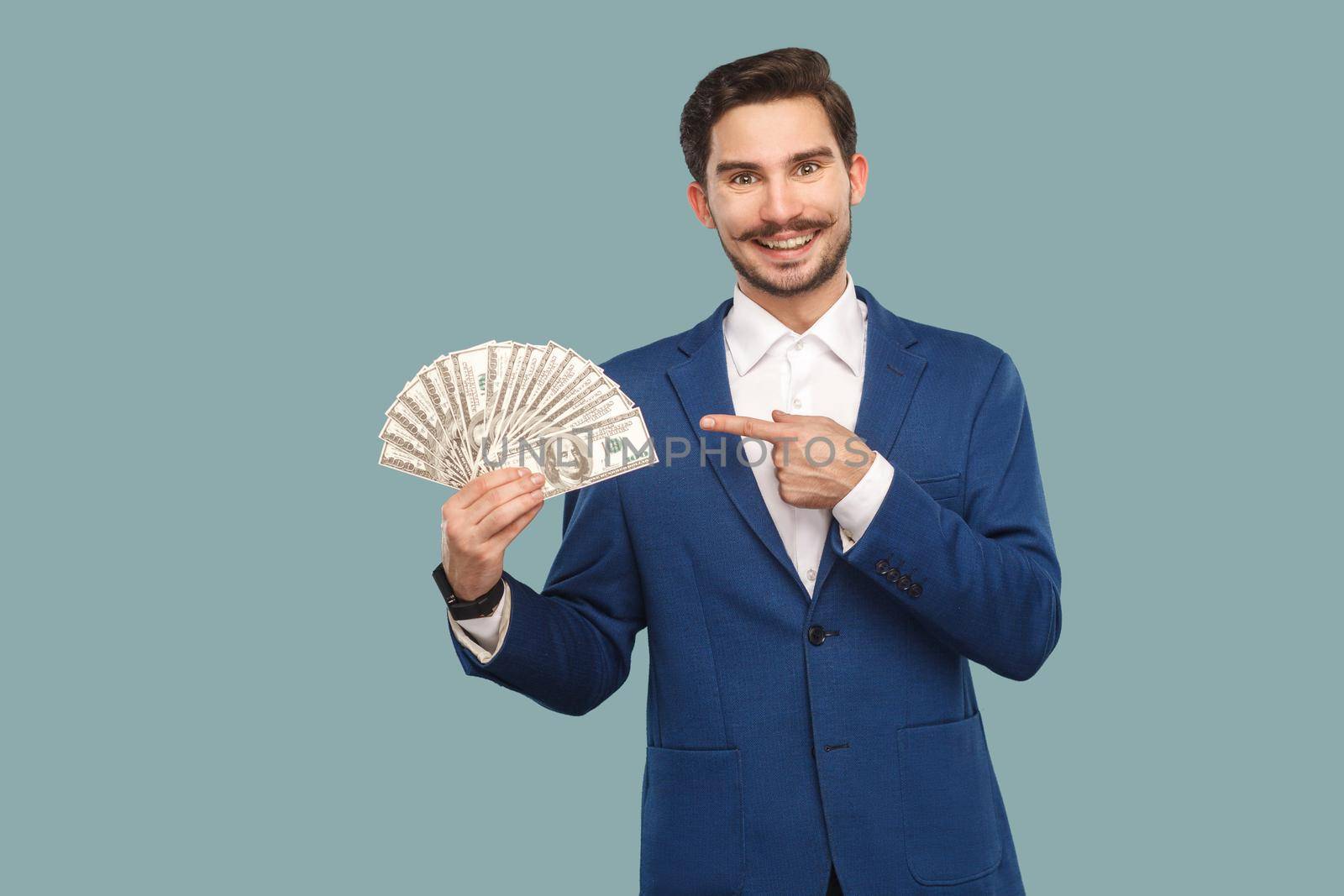 Handsome happy businessman in blue jacket standing and holding many dollars in hand and looking at camera with pointing finger and toothy smile . Indoor, studio shot isolated on light blue background