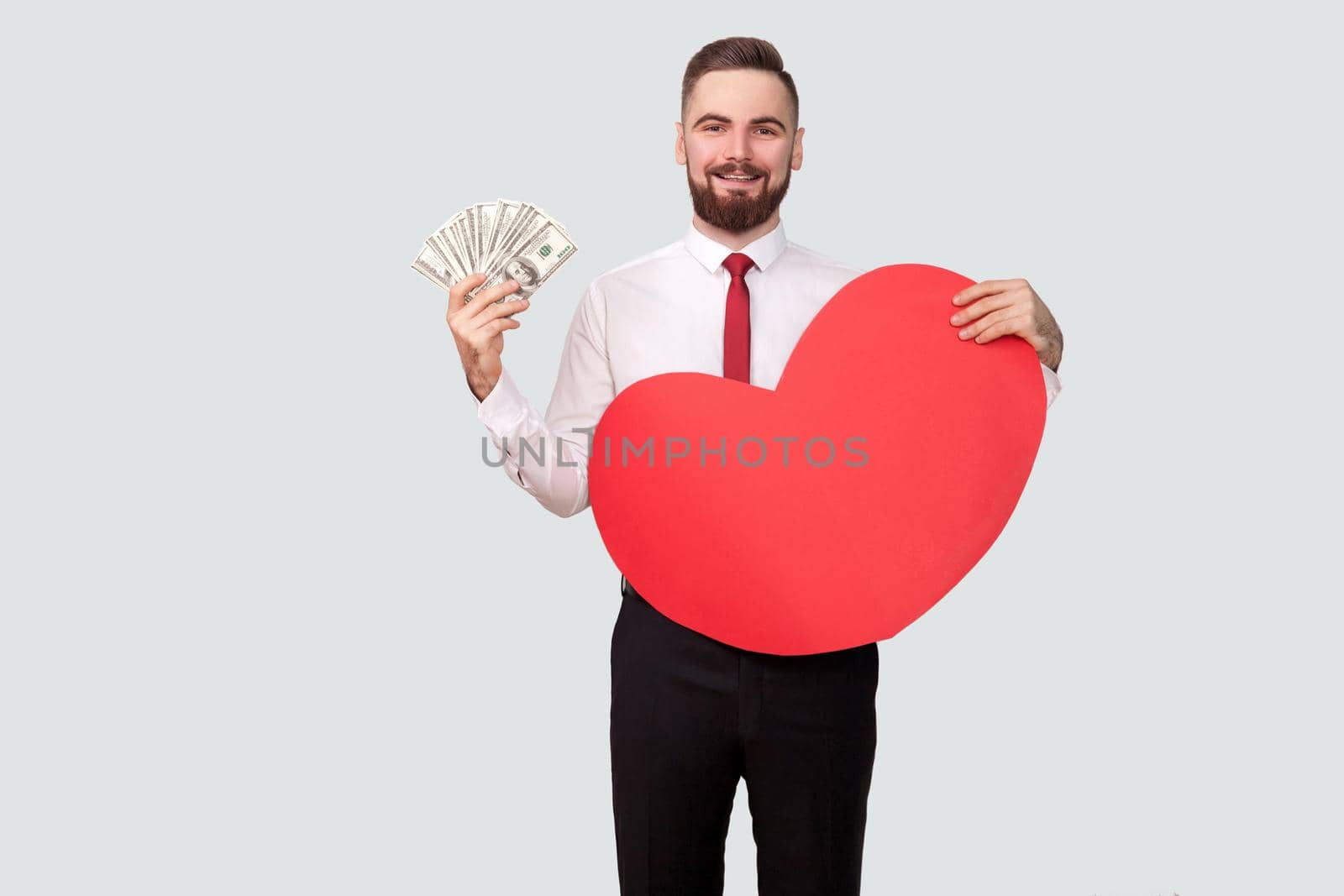 Young bearded man holding many dollars and red heart shape and looking at camera with toothy smile. Indoor, studio shot, isolated on gray background