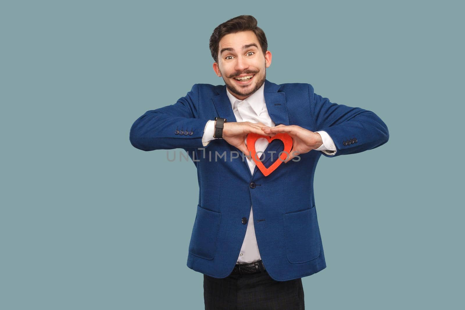 Handsome man in blue jacket and white shirt, standing and holding red heart shape and looking at camera with toothy smile. Indoor, studio shot isolated on light blue background