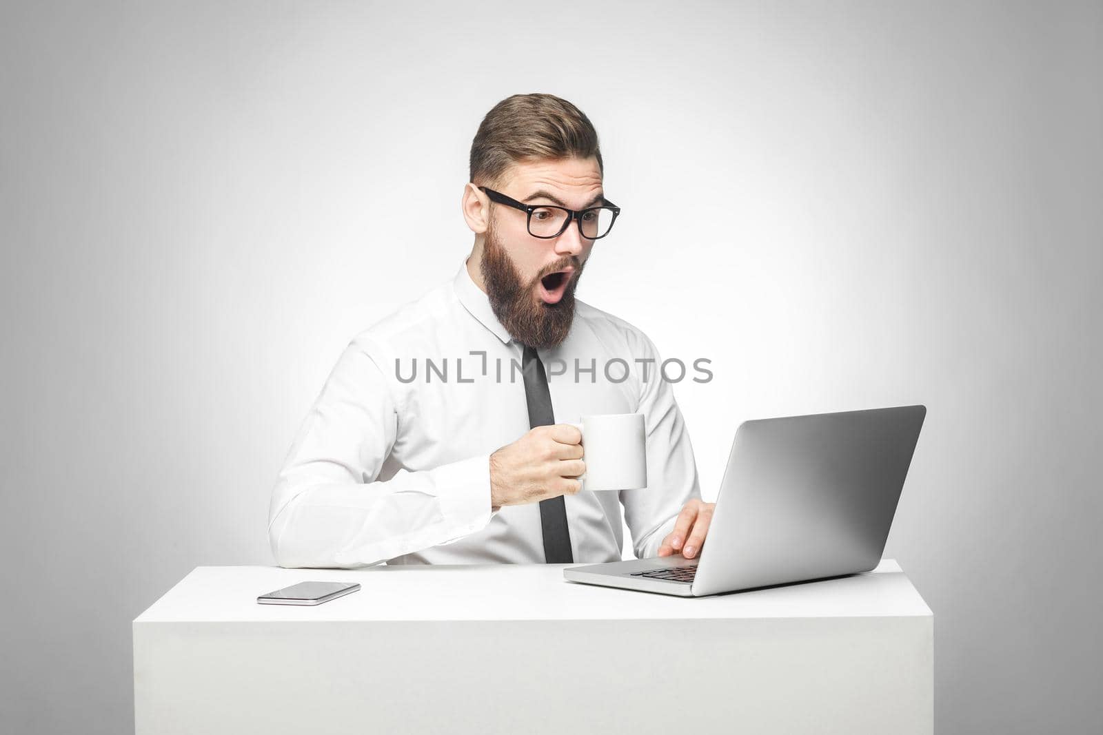 Can't believe! Portrait of emotional shocked young businessman in white shirt and black tie are sitting in office, reading news and remotely working with suprised big eyes looking at laptop. Isolated