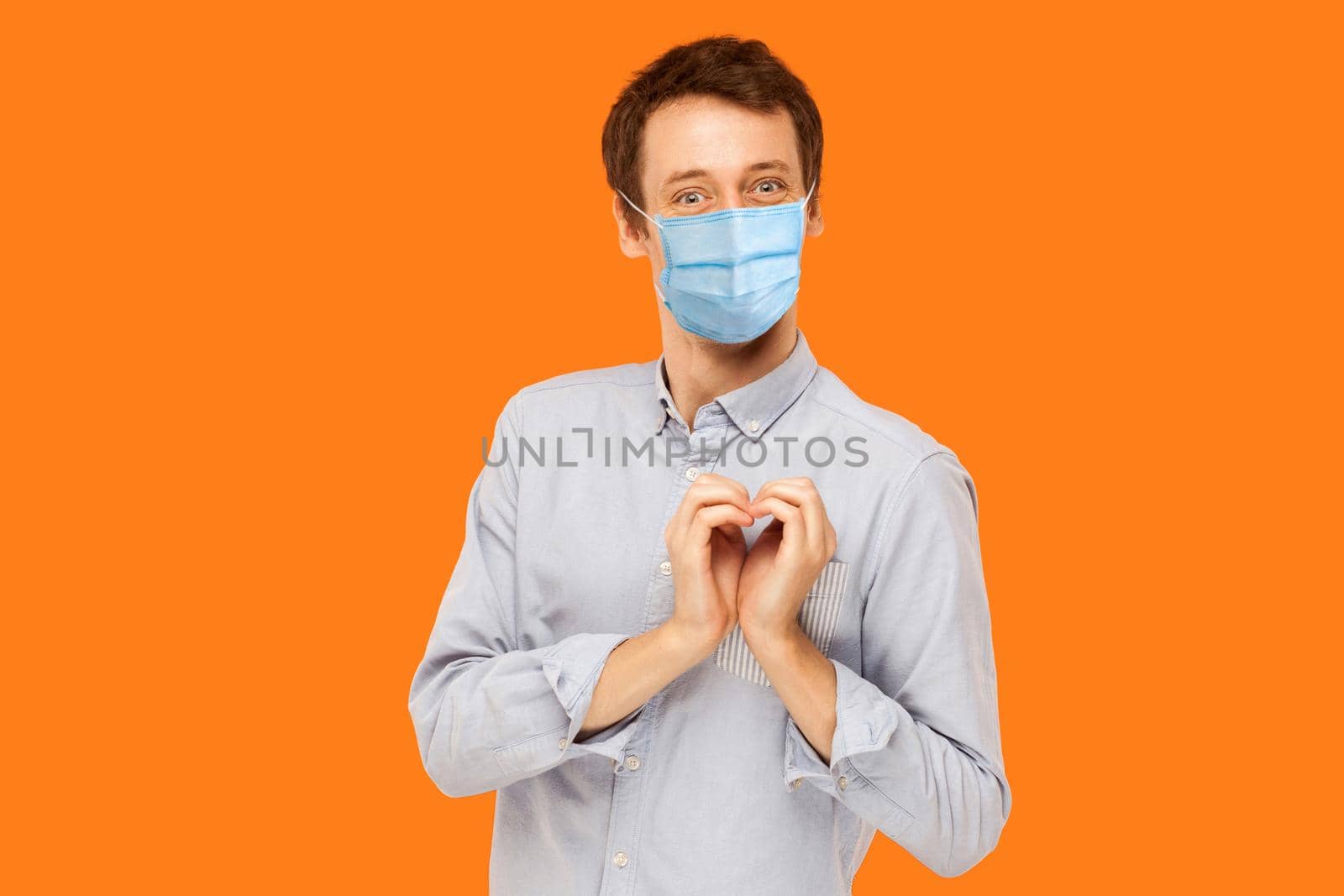 I love you. Portrait of joyful happy young worker man with surgical medical mask standing with heart love gesture and looking at camera with smile. indoor studio shot isolated on orange background.