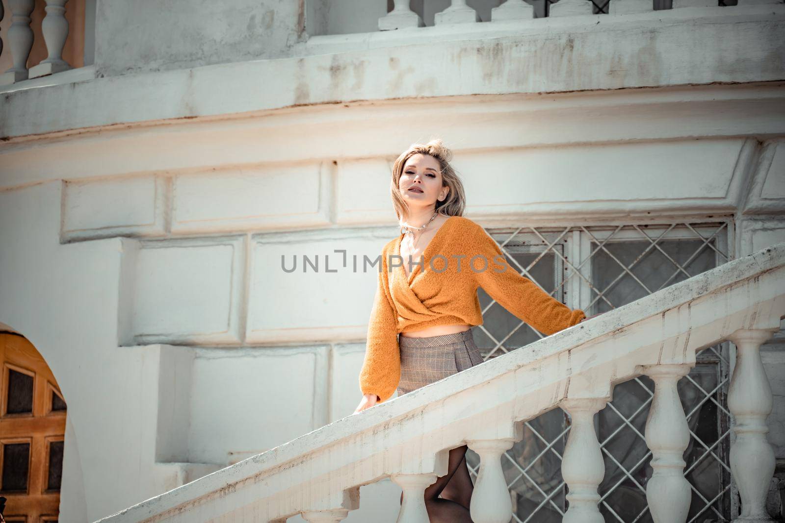 A middle-aged woman looks like a good blonde with curly beautiful hair and makeup on the background of the building. She is wearing a yellow sweater