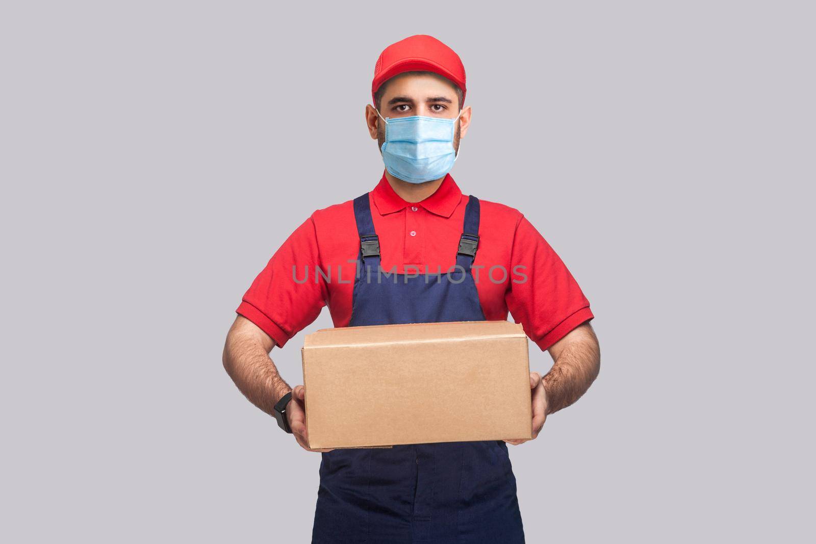 Delivery on quarantine. Portrait of young man with surgical medical mask in blue uniform and red t-shirt standing and holding the cardboard box on grey background. Indoor, studio shot, isolated,