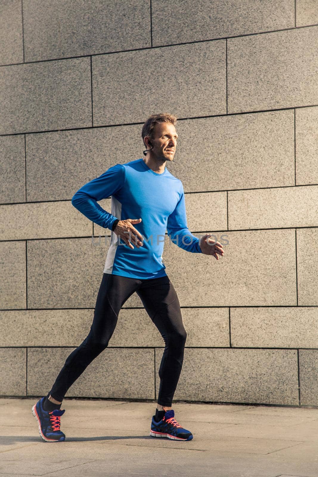 Handsome middle aged serious man in blue black sports uniform and headphones and fitness tracker is running in the city.