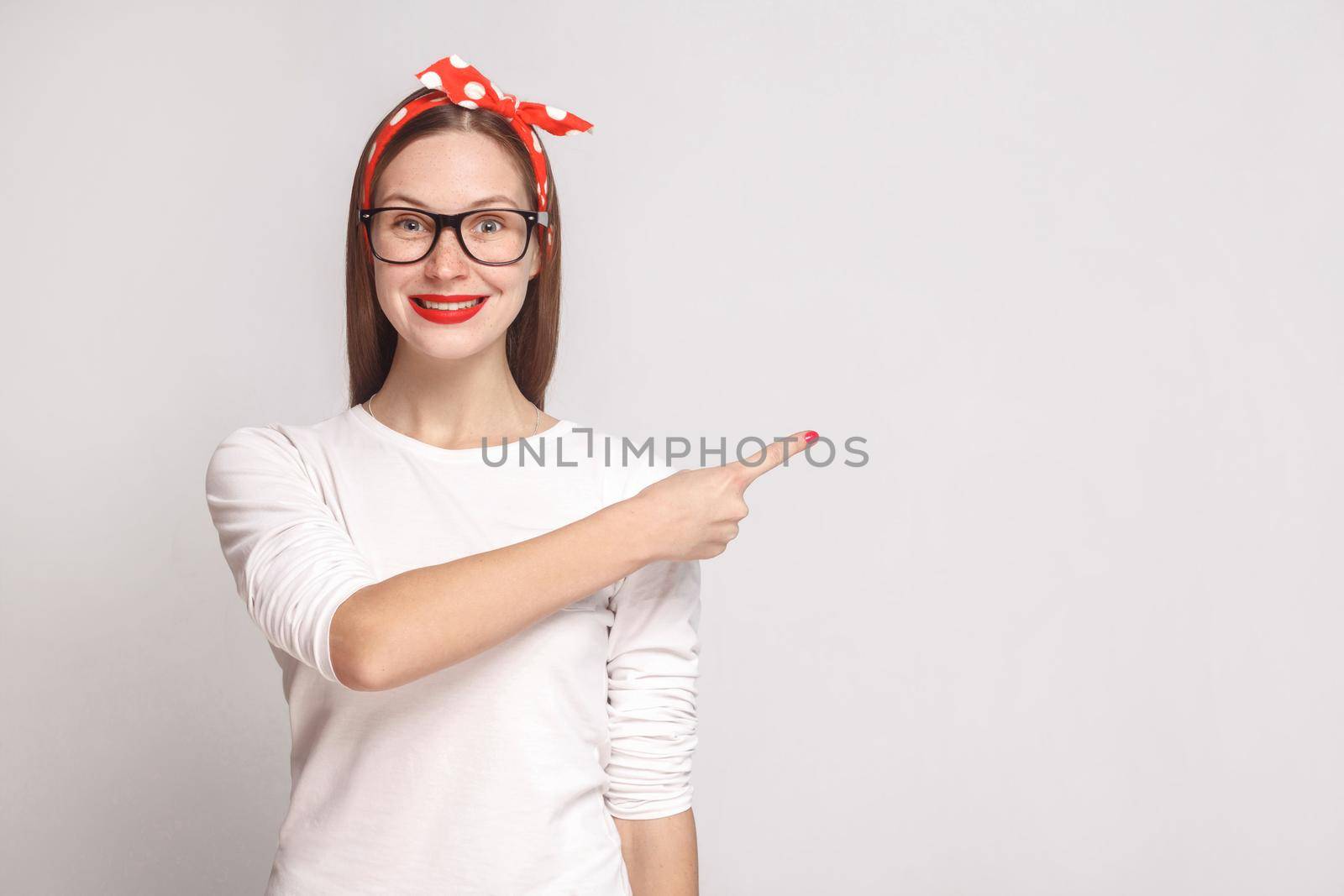 look at this side. pointing finger. portrait of beautiful emotional young woman in white t-shirt with freckles, glasses, red lips and head band. indoor studio shot, isolated on light gray background.