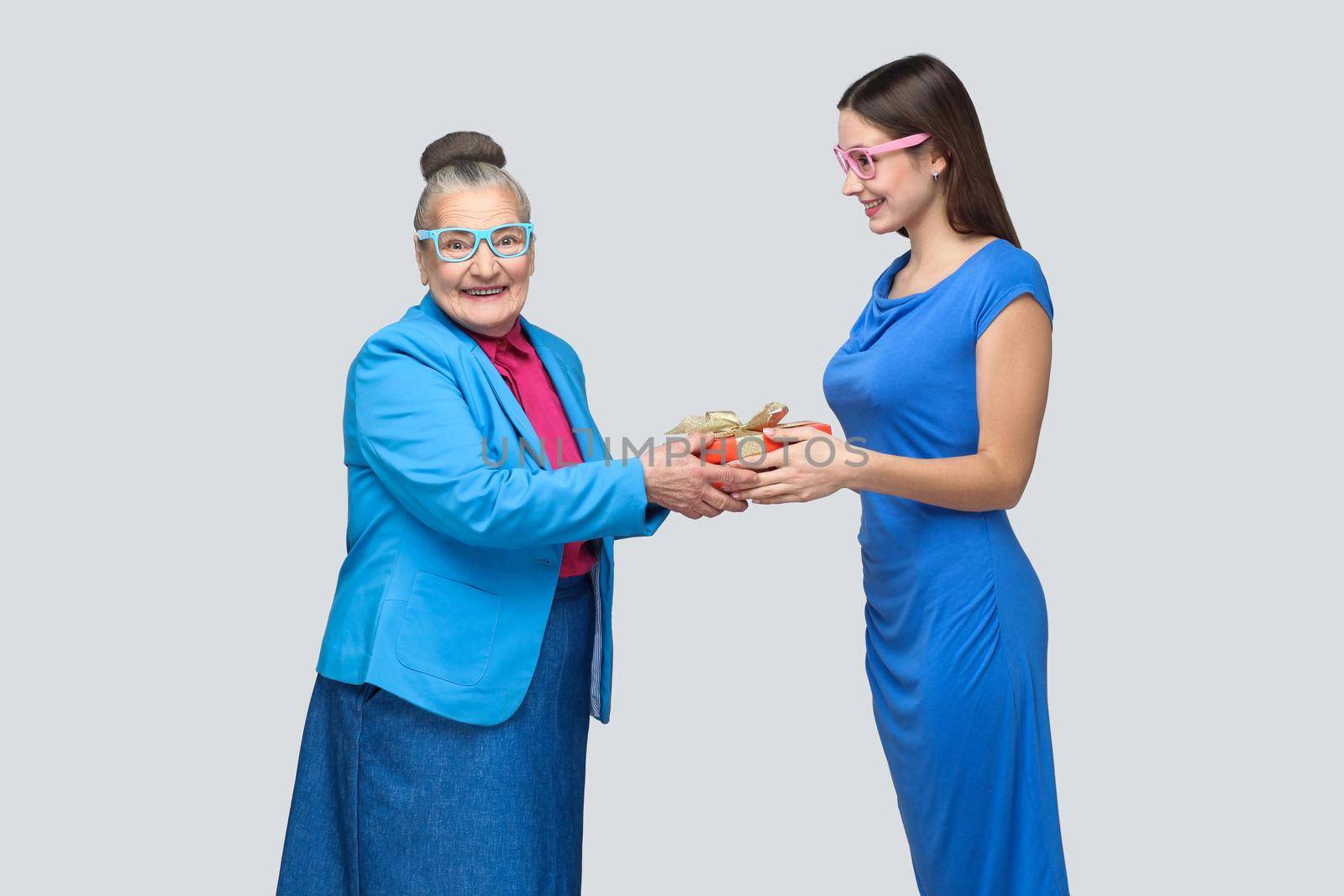 Granddaughter in blue dress smiles and gives a gift to her lovely grandmother, looking at camera with surprised face. Friendship mutual understanding. indoor, studio shot, isolated on gray background.