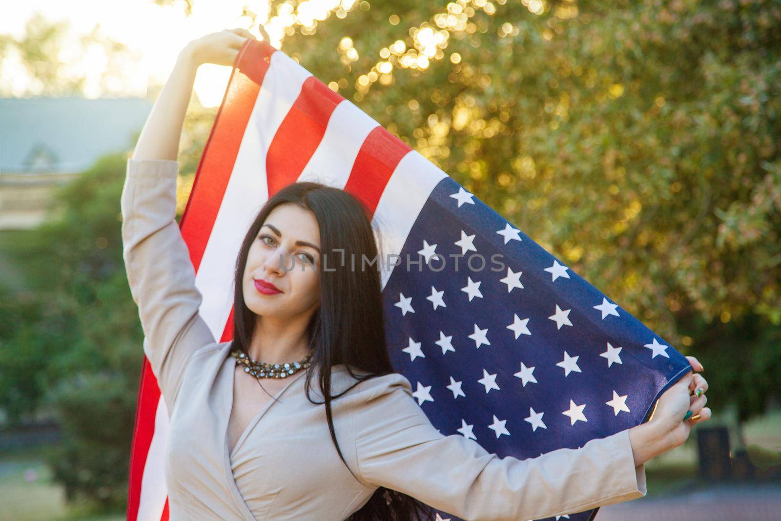 American flag and woman (4th july). Beautiful young woman with classic dress holding american flag in the park. fashion model holding us smiling and looking at camera. usa lifestyle..