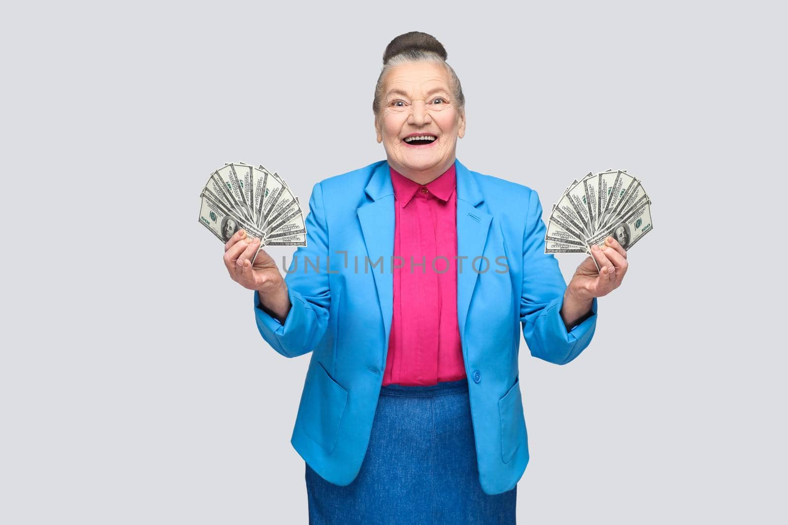 happy stylish aged woman holding many american dollars. Portrait of handsome expressive grandmother in light blue suit with collected gray hair bun hairstyle. Studio shot, isolated on gray background