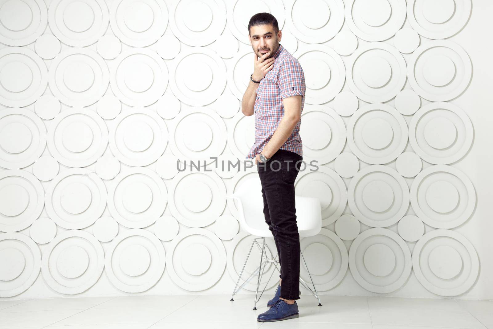 Young bearded fashion model in casual style is posing near white circle wall background. studio shot. looking at camera with smile.