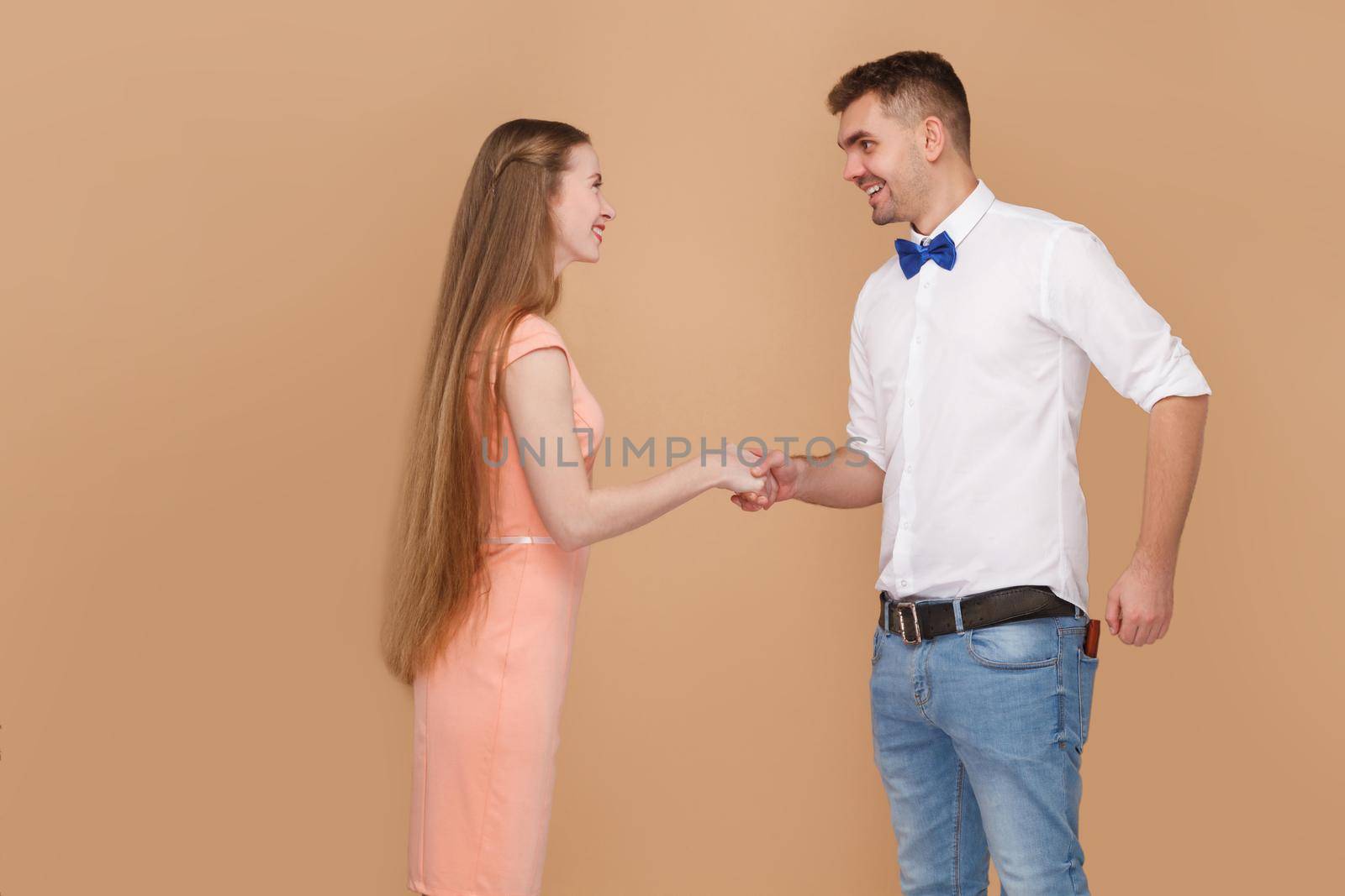 Nice to meet you. Profile side view handshake of handsome man in white shirt and long hair blonde woman in pink dress looking with toothy smile. indoor studio shot, isolated on light brown background