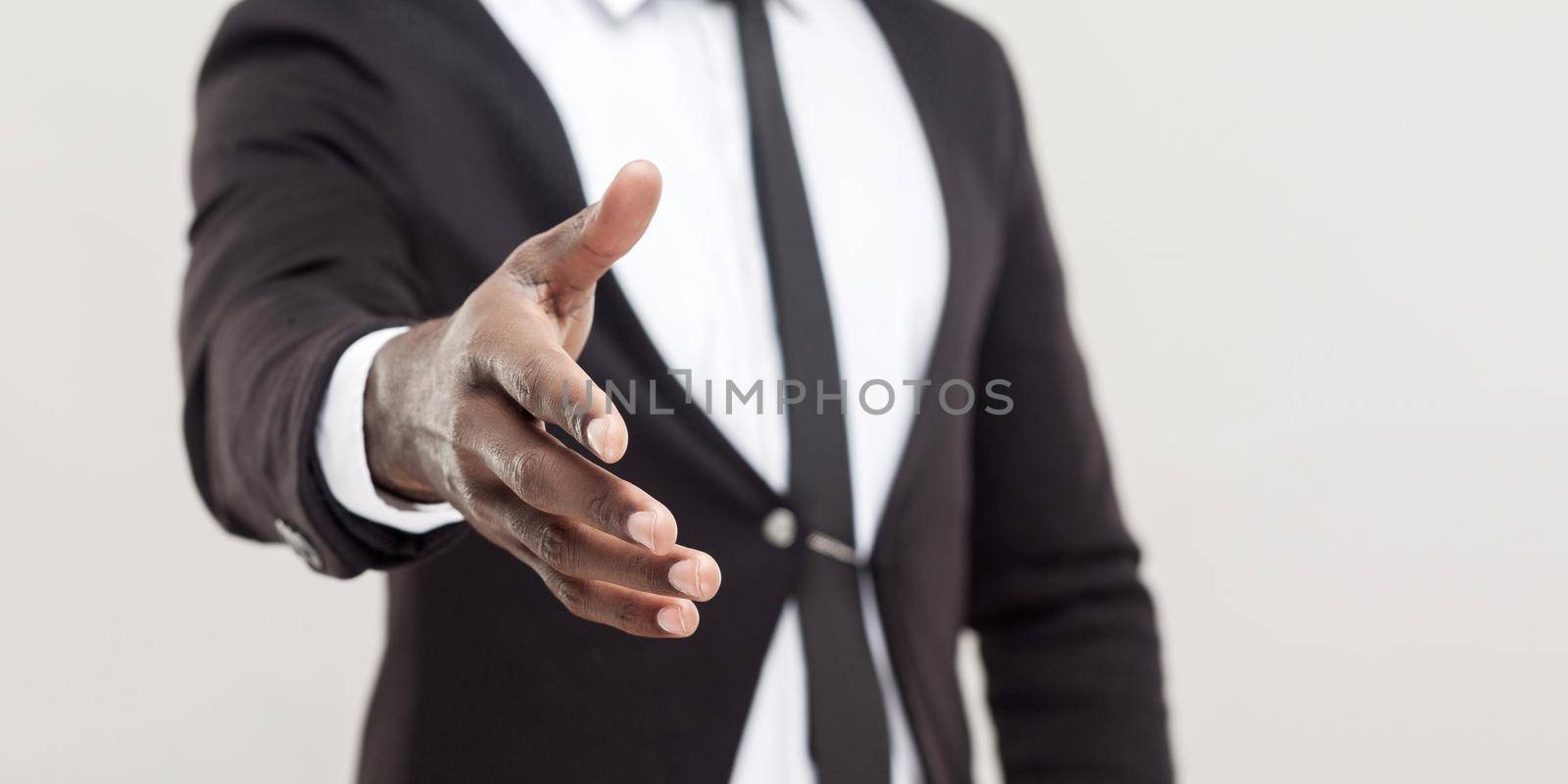 Man hand in black suit and tie giving hand to greeting or handshake at camera. focus on hand. indoor studio shot, isolated on light grey background copyspace.