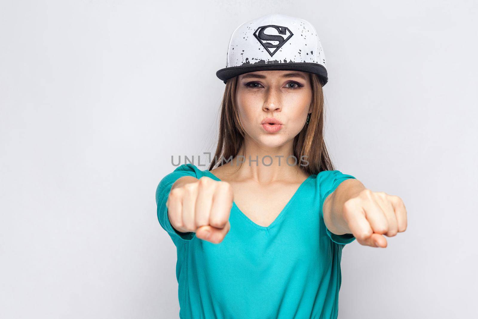 Portrait of cool dangerous beautiful young girl in blue dress and white cap with supermen sign standing, showing dukes and ask left or right. Indoor, studio shot, copy space, grey background