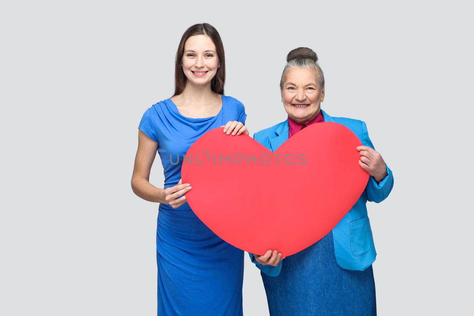 happy granddaughter and grandmother standing, holding big heart shape, looking at camera with toothy smile. Big love in family. Relations in the family. indoor studio shot, isolated on gray background