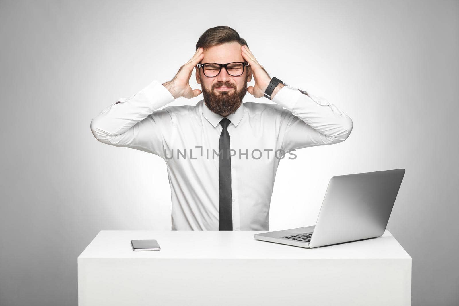 Headache. Portrait of unhealphy painful young manager in white shirt and black tie are sitting in office and have strong headache, holding fingers near temples. Studio shot, isolated, gray background