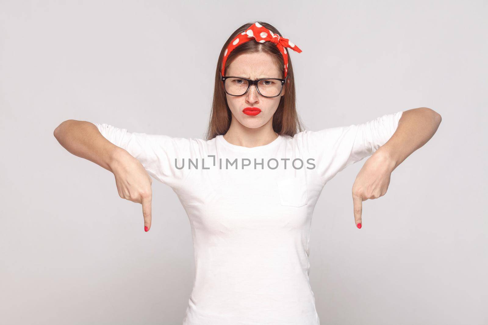 must be here right now. bossy anger portrait of beautiful emotional young woman in white t-shirt with freckles, glasses, red lips and head band. indoor studio shot, isolated on light gray background.